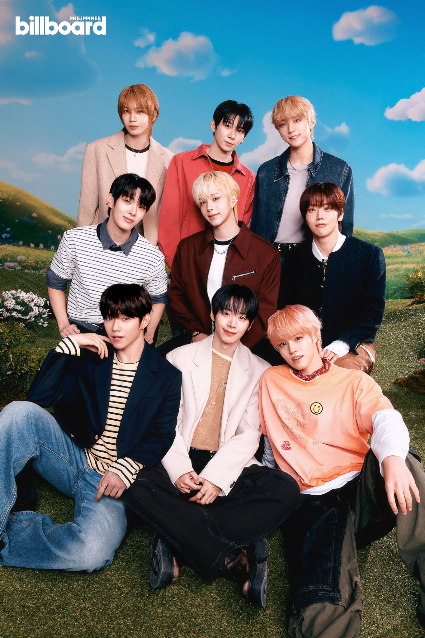 Seven members of a boy band posing together in a sunny outdoor field with a blue sky backdrop and fluffy clouds in the background.