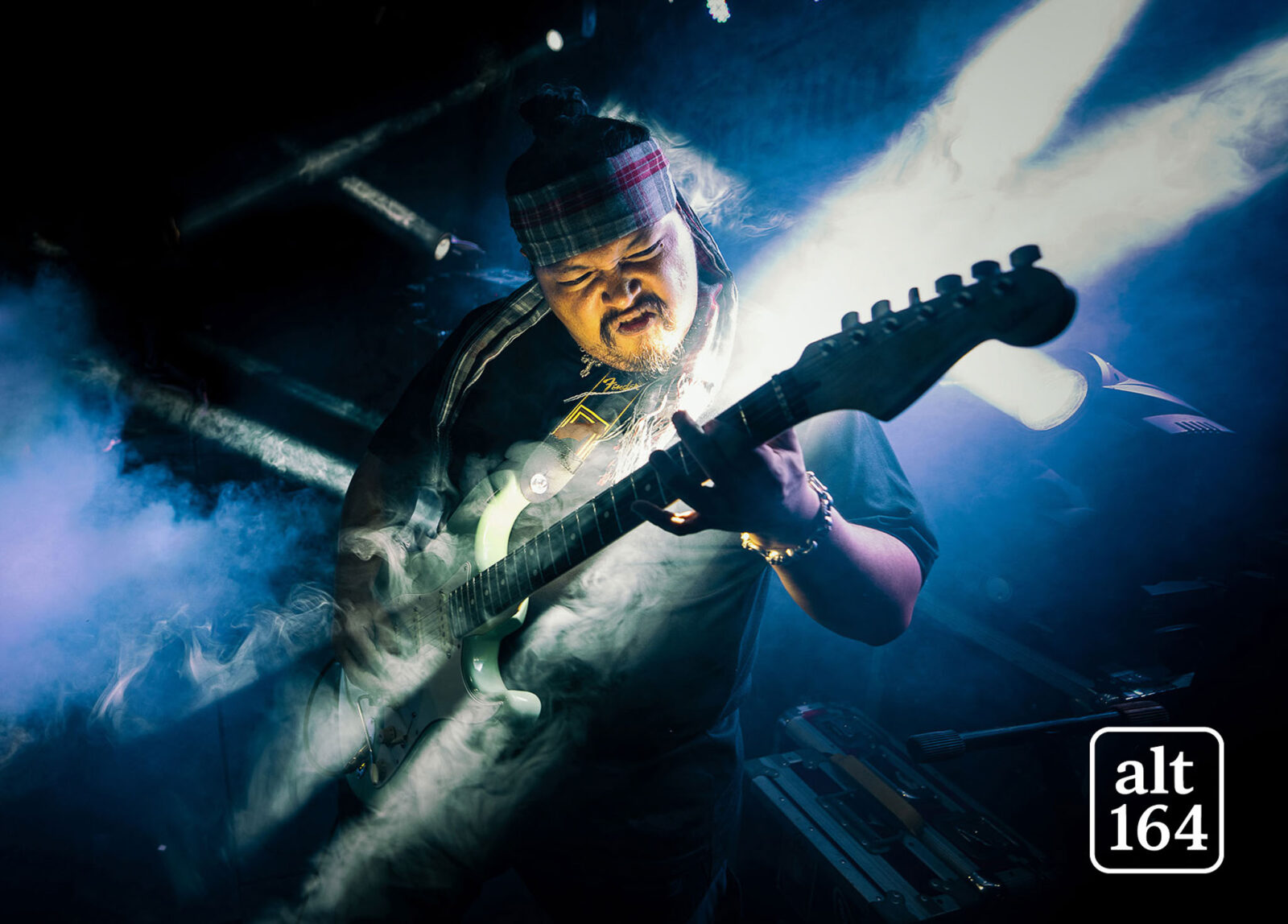 Guitarist wearing a plaid headband and scarf plays a white electric guitar amid blue stage lights and smoke.