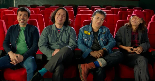 Four men sit in red theater seats, smiling at the camera in a movie theater.