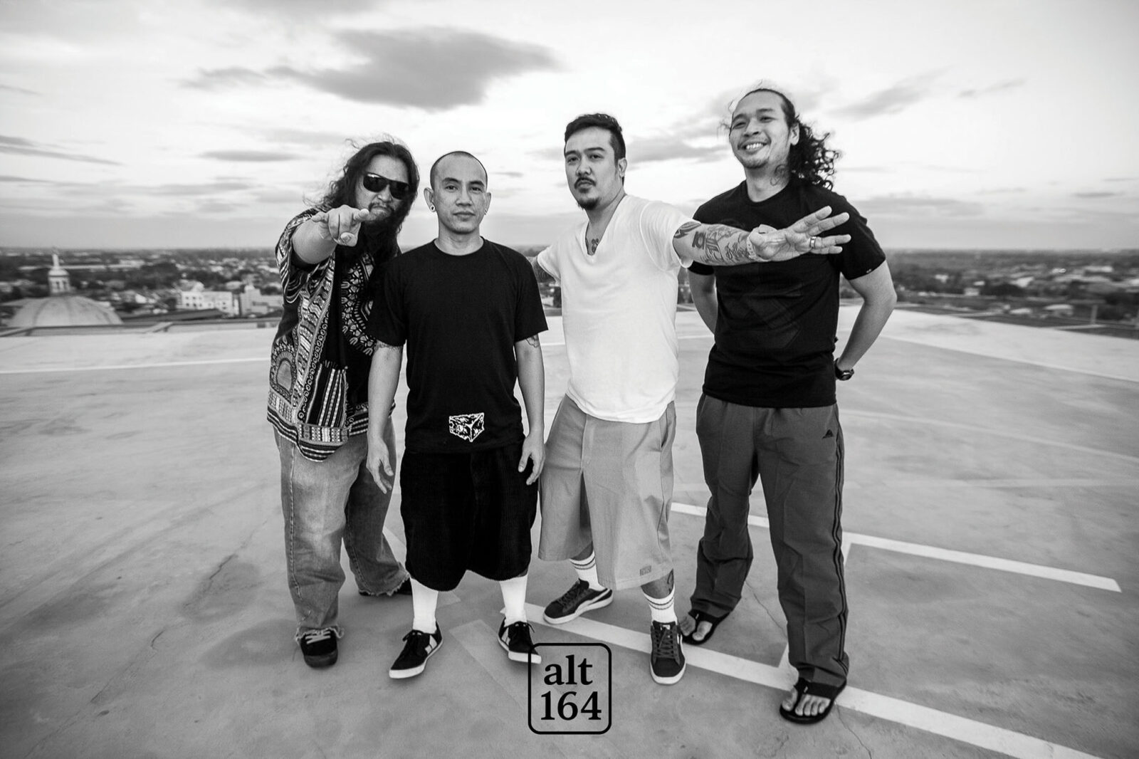 Four men stand together on a rooftop, one pointing at the camera, with a cityscape in the distance (black-and-white photo).