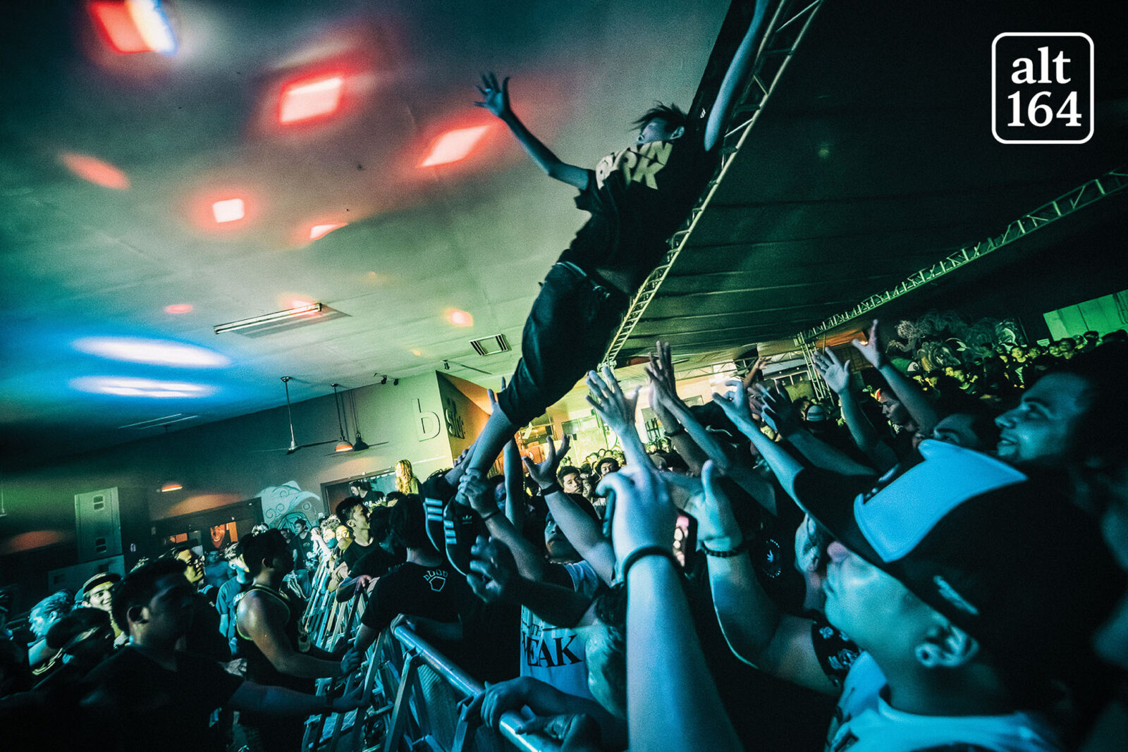 Person crowd-surfing over the barrier at a packed indoor concert, arms raised and a sea of fans cheering below, colorful stage lights overhead