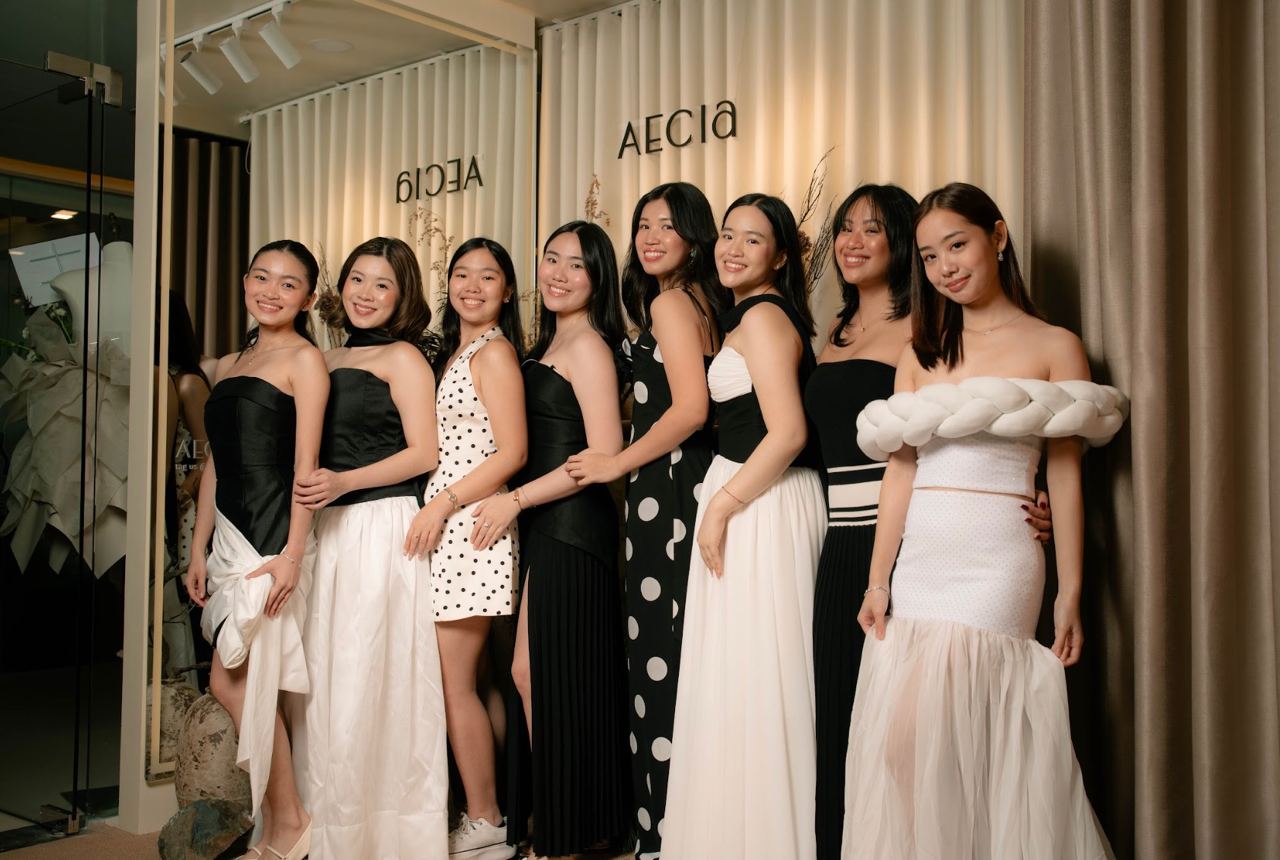 Nine women in elegant black-and-white dresses pose together at a fashion event, smiling in front of a beige curtain backdrop with'AECIA' signage.