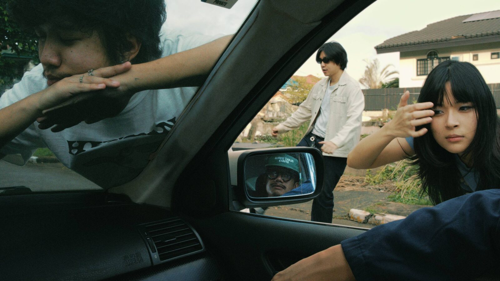 Three people lean into a car window from outside: a woman with long hair on the right, a man in a light jacket on the left, and a man leaning in from the driver's side with a white shirt visible in the background.