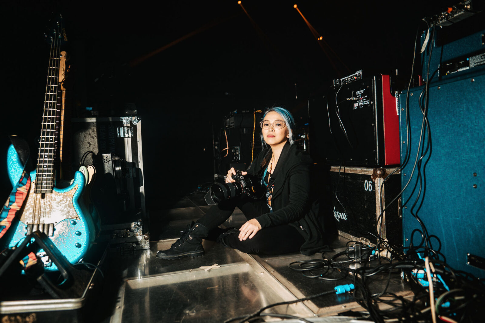 Female photographer with a camera sits on the backstage floor among gear and cables, a blue guitar to her left and dim stage lights in the background.