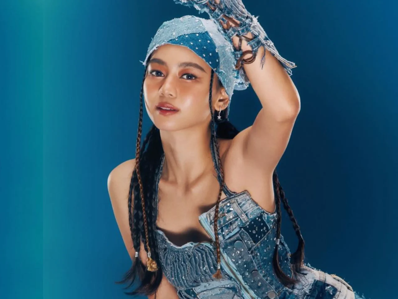 Woman model in a blue denim outfit poses with one arm raised against a teal-blue gradient background.