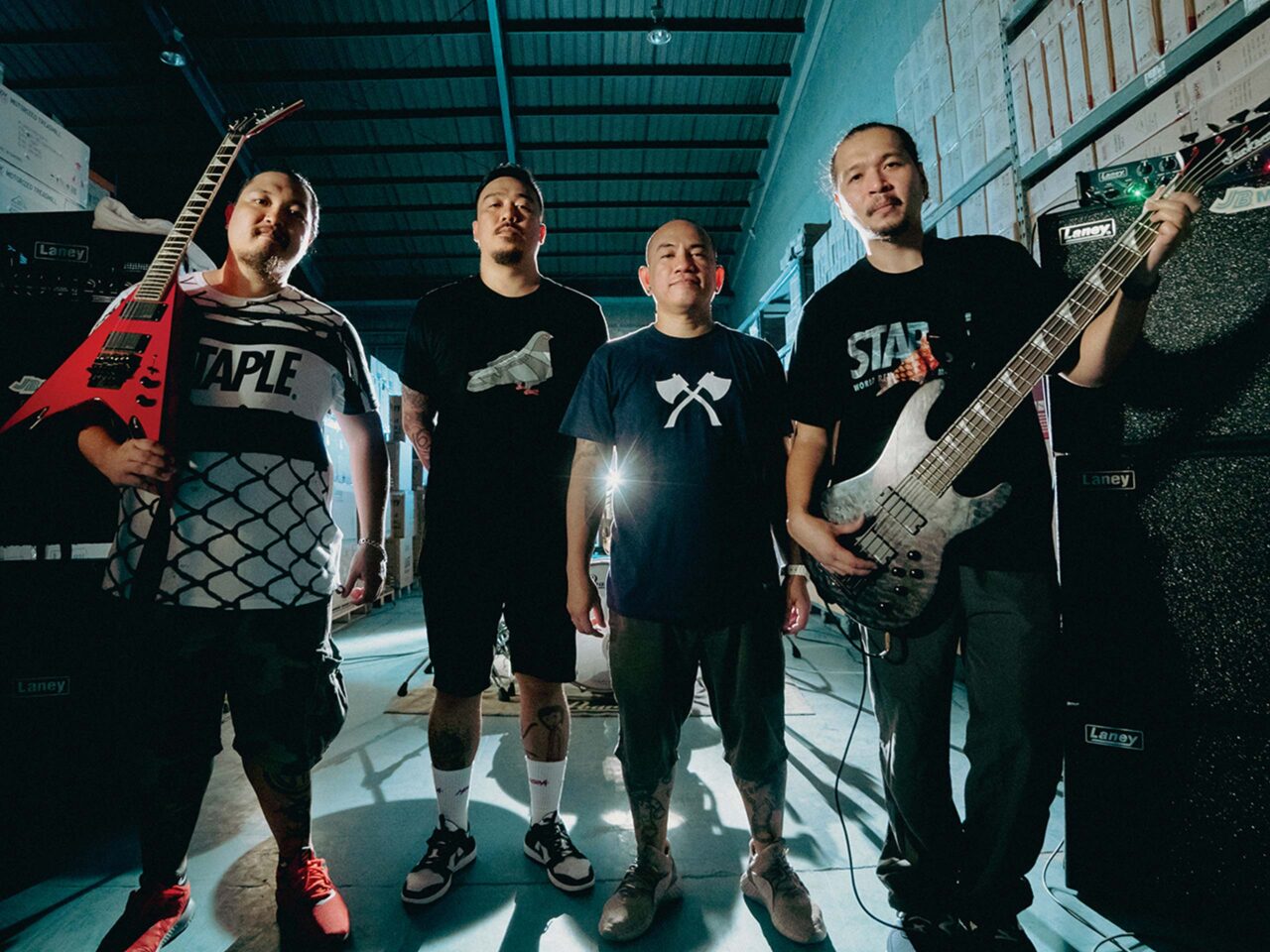 Four musicians stand in a backstage warehouse with amplifiers; two hold guitars/bass.