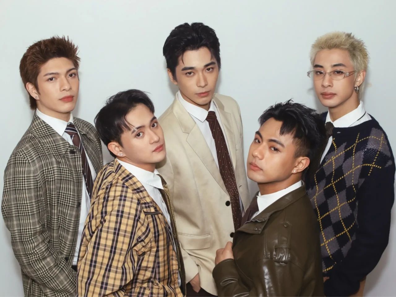 Six young men posing together in stylish suits and jackets against a light backdrop.