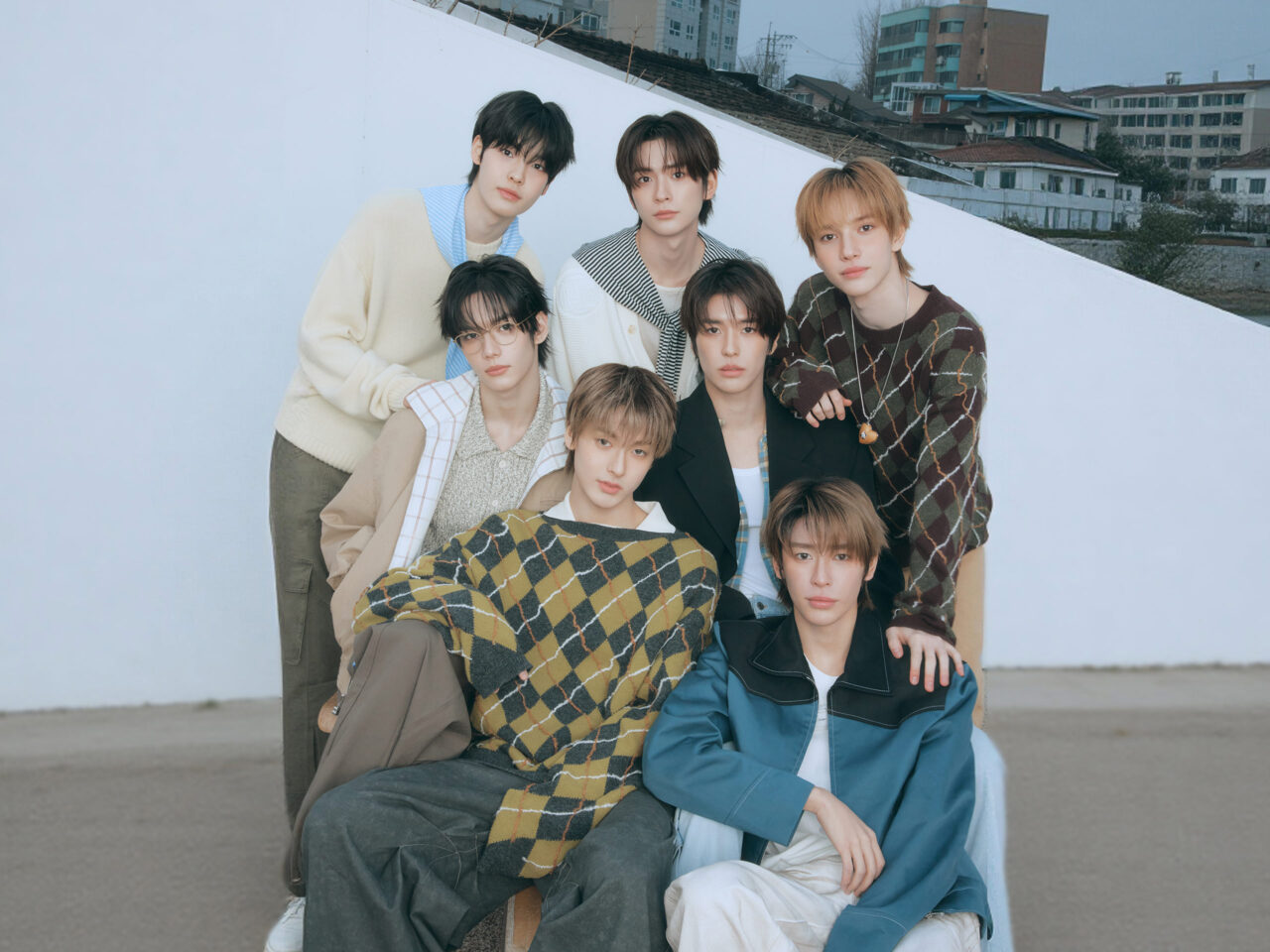 Seven young men posing together outdoors in stylish outfits against a pale blue wall backdrop.
