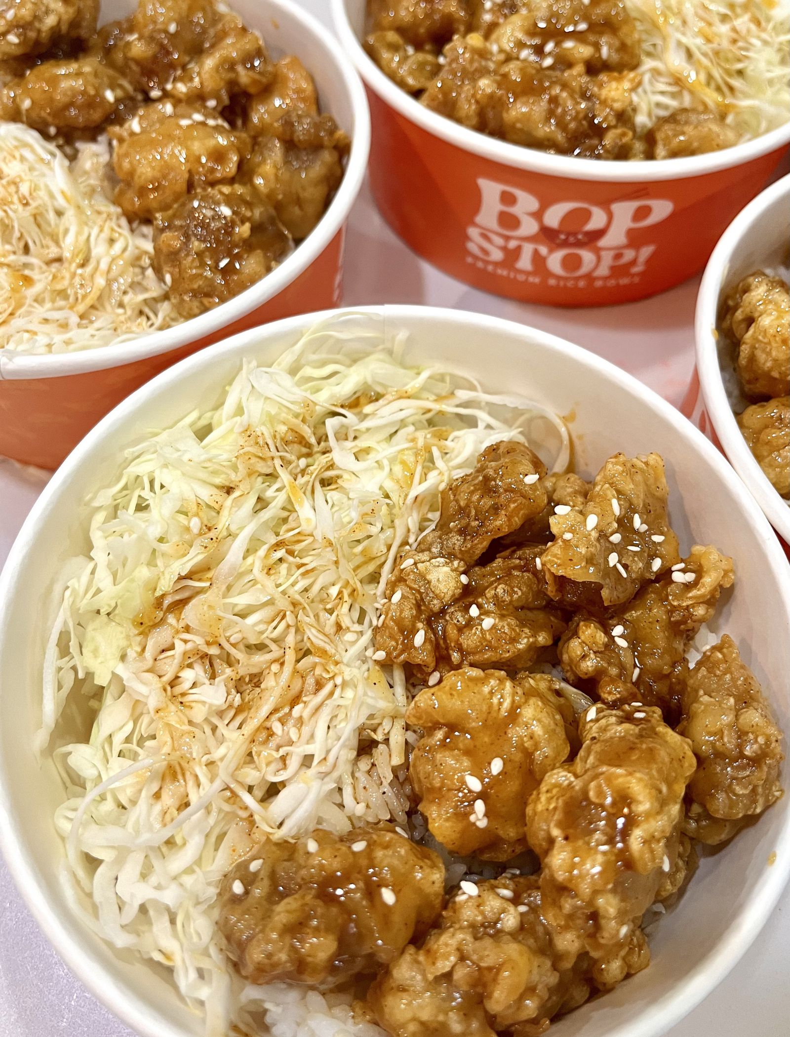 Bowl of glazed fried chicken pieces with sesame seeds served over white rice and shredded cabbage in an orange container.