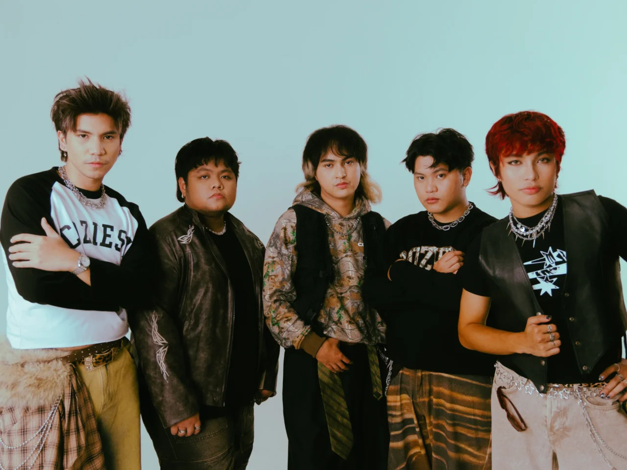 Five young men posing together for a photo in trendy streetwear against a light teal background, showing confident expressions and layered outfits.