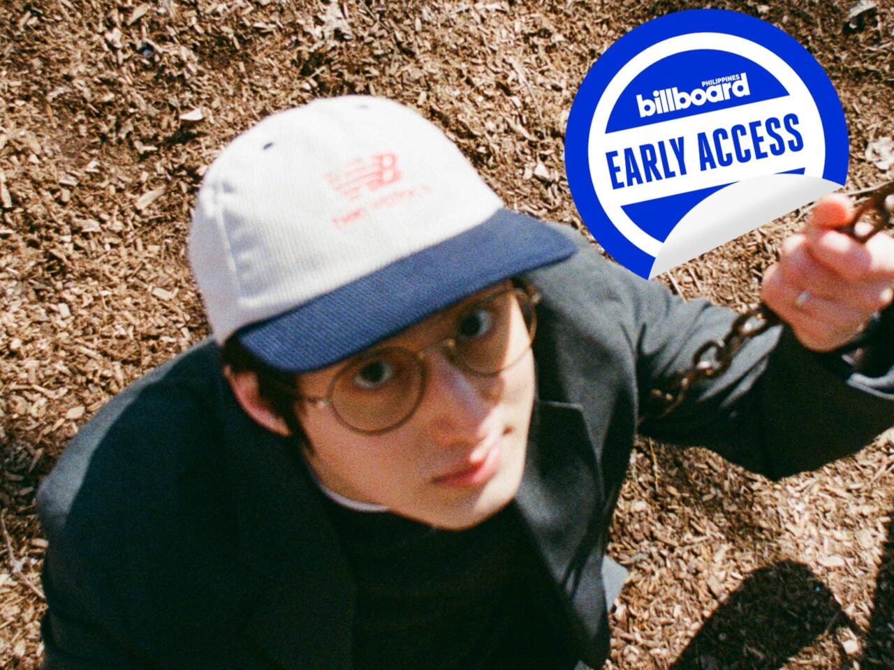 Person wearing glasses and a white cap reaches toward the camera while holding a swing chain, on a wood-chip playground with a peeling Billboard Early Access sticker in the left foreground.