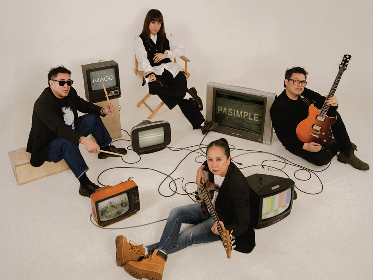 Four musicians pose in a white studio surrounded by vintage CRT televisions, cables, and their instruments (guitars and microphone).