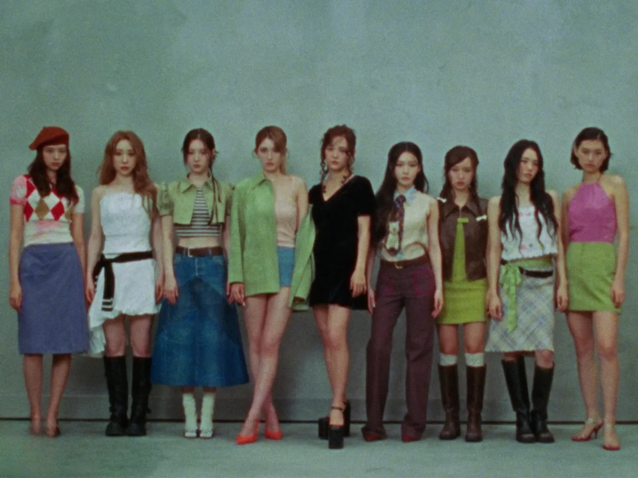 Group of ten women standing in a row against a plain wall, wearing colorful retro outfits and boots.