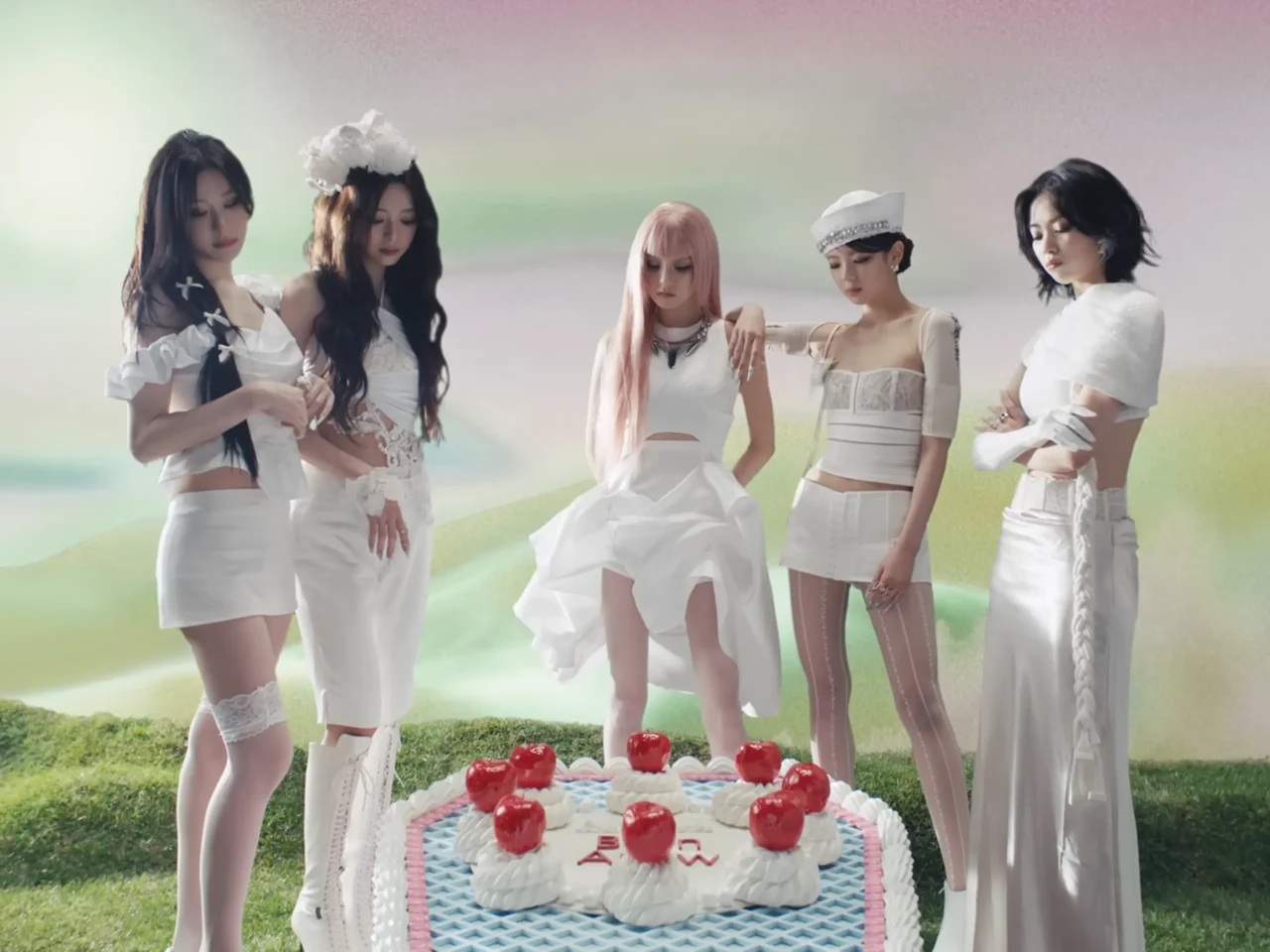 Six women dressed in white outfits pose around a white frosted cake with red cherries on a grassy landscape backdrop.