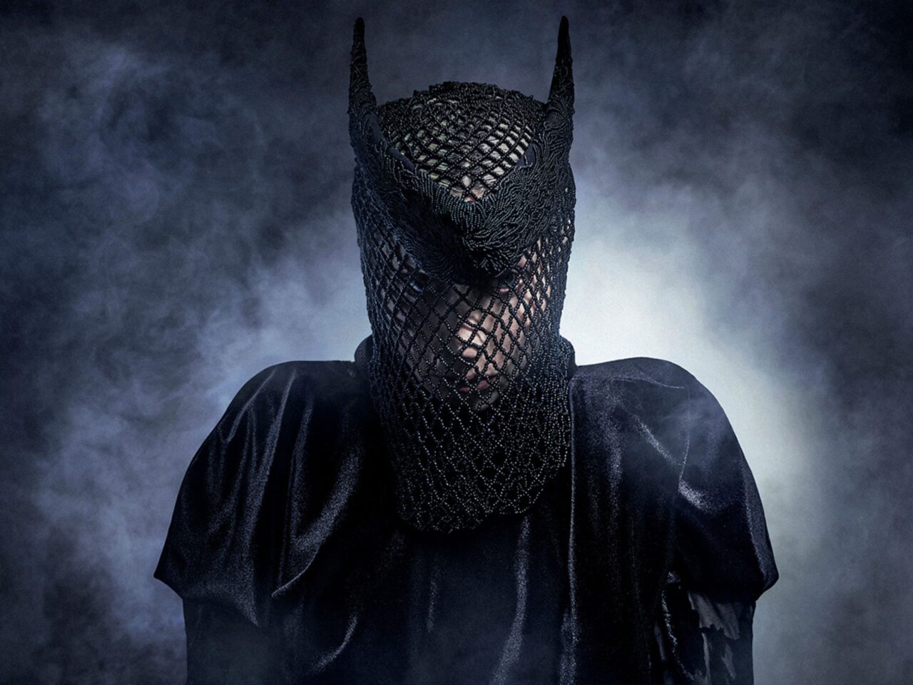 Person wearing a black chainmail hood that covers the head and neck, standing in a misty blue background.