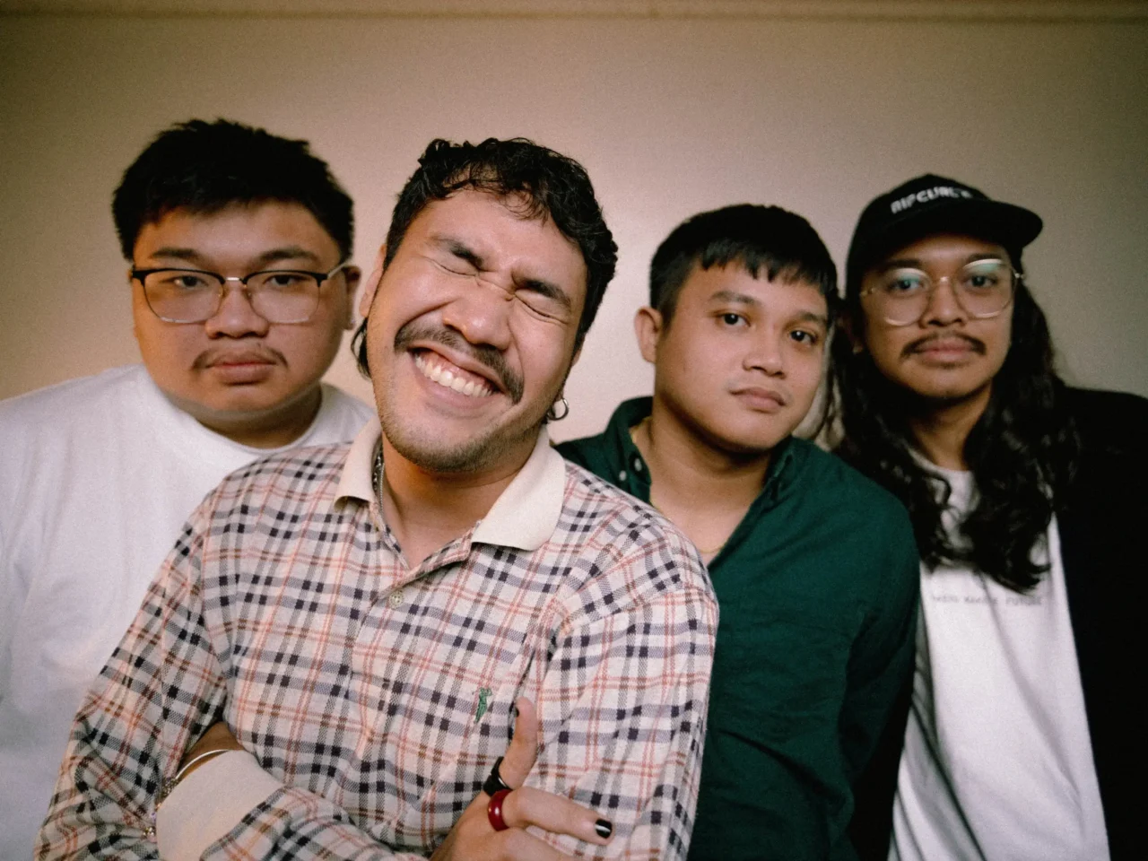 Four friends posing closely for a casual group photo, with one man in plaid smiling broadly in the foreground.