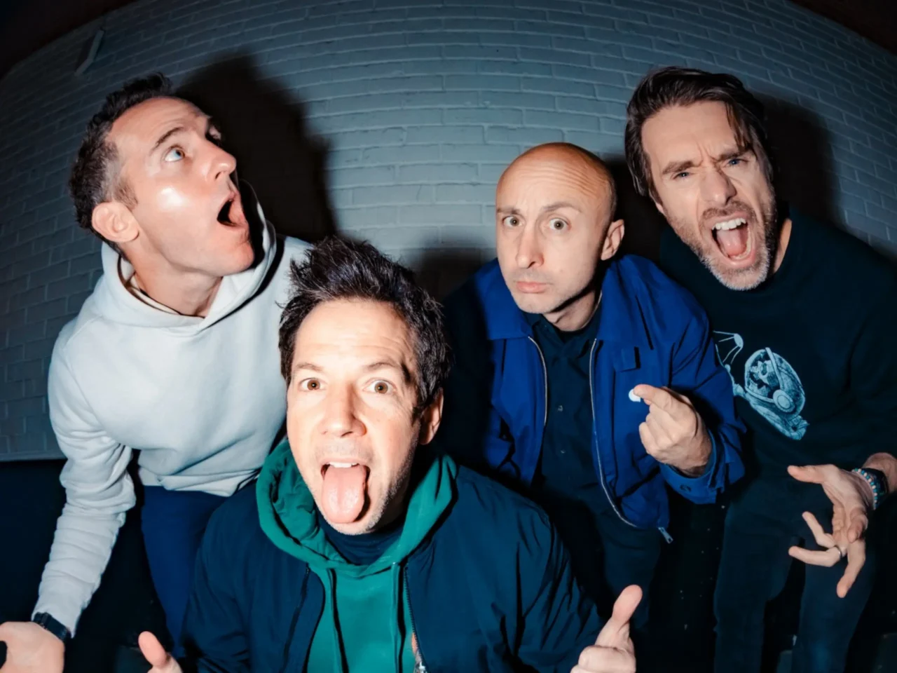 Four men in casual jackets lean toward the camera, making goofy faces and sticking out their tongues against a blue brick wall.
