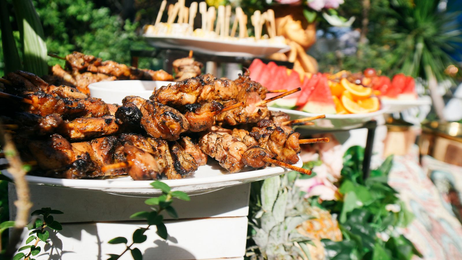 Skewers of char-grred grilled meat piled on a platter at an outdoor buffet, with fruit platters in the background.