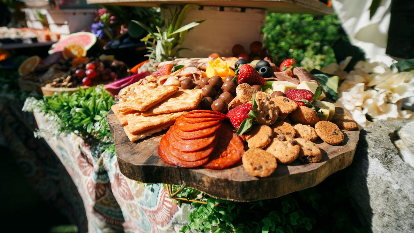 Outdoor charcuterie spread on a rustic wooden board: crackers, salami, cheeses, berries, cookies, and chocolate treats, with greenery in the background.