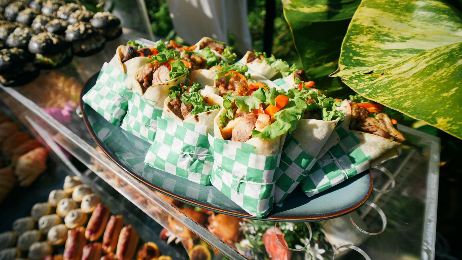 Plate of wrap sandwiches filled with grilled chicken, lettuce, and peppers on green-and-white checkered wrappers outdoors.