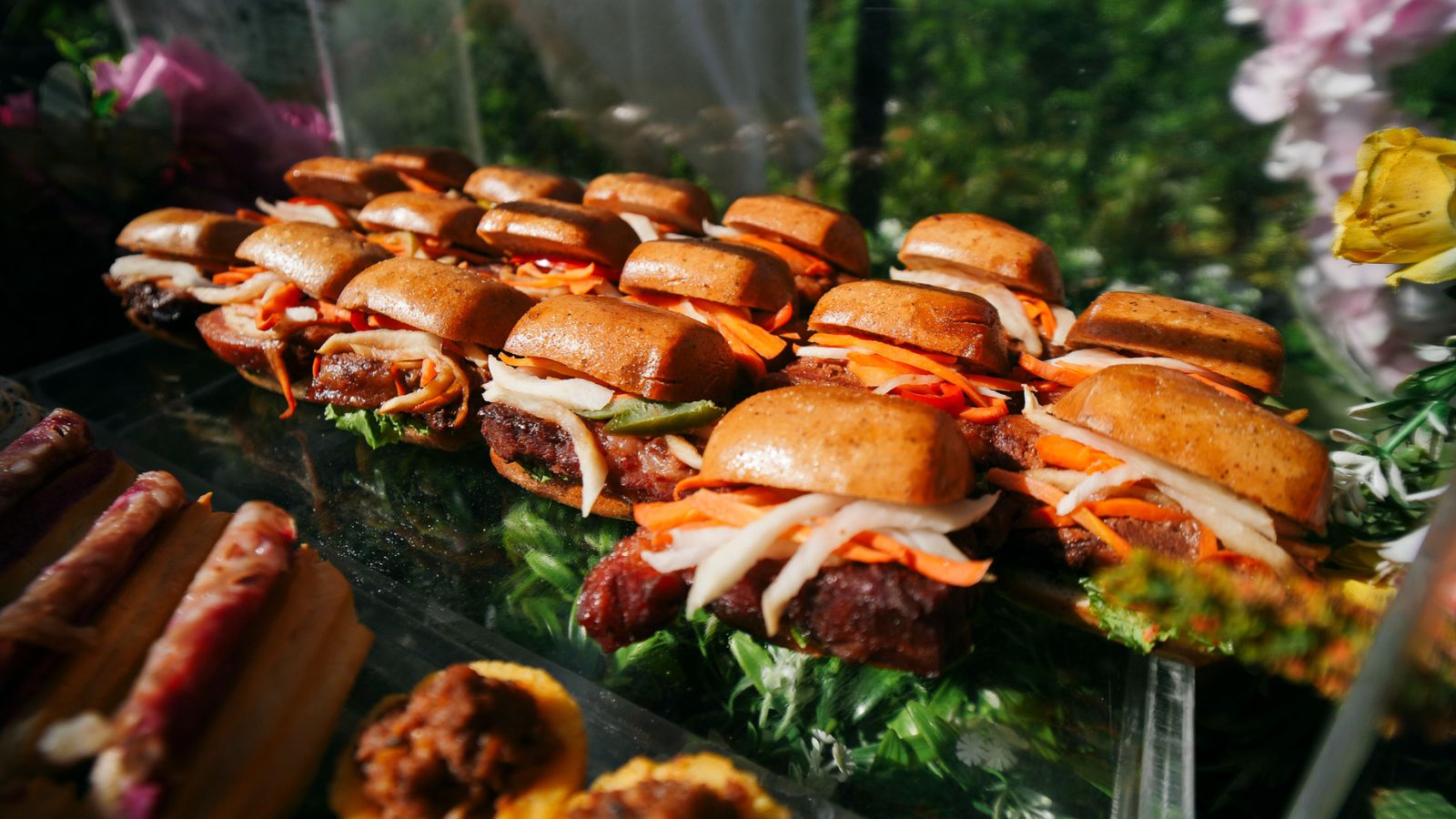 Row of mini beef sliders with carrot slaw on glossy buns arranged on a display tray.