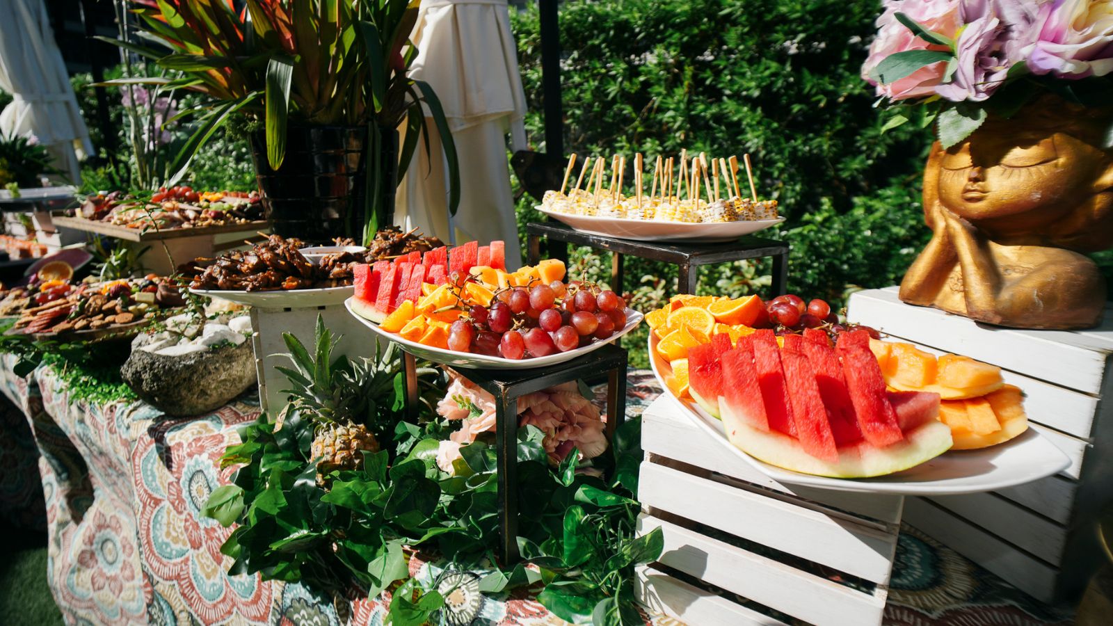 Outdoor garden buffet with colorful fruit platters (watermelon, grapes, cantaloupe) and skewered bites on a sunny table.