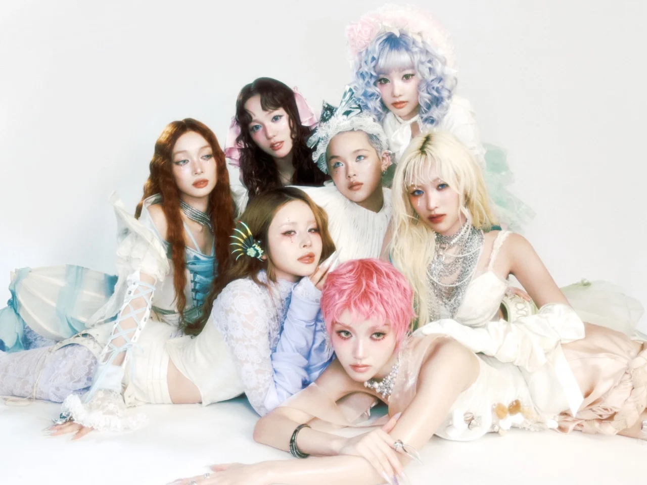 Group portrait of seven young women in pastel, fantasy-style outfits and colorful hair against a pale background, posing closely together.