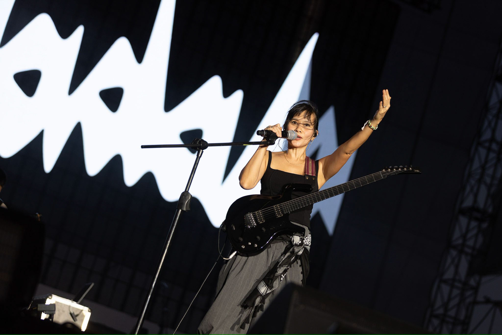 Female singer with a black electric guitar performing on stage, mic in hand, one arm raised in the air against a large black-and-white backdrop.