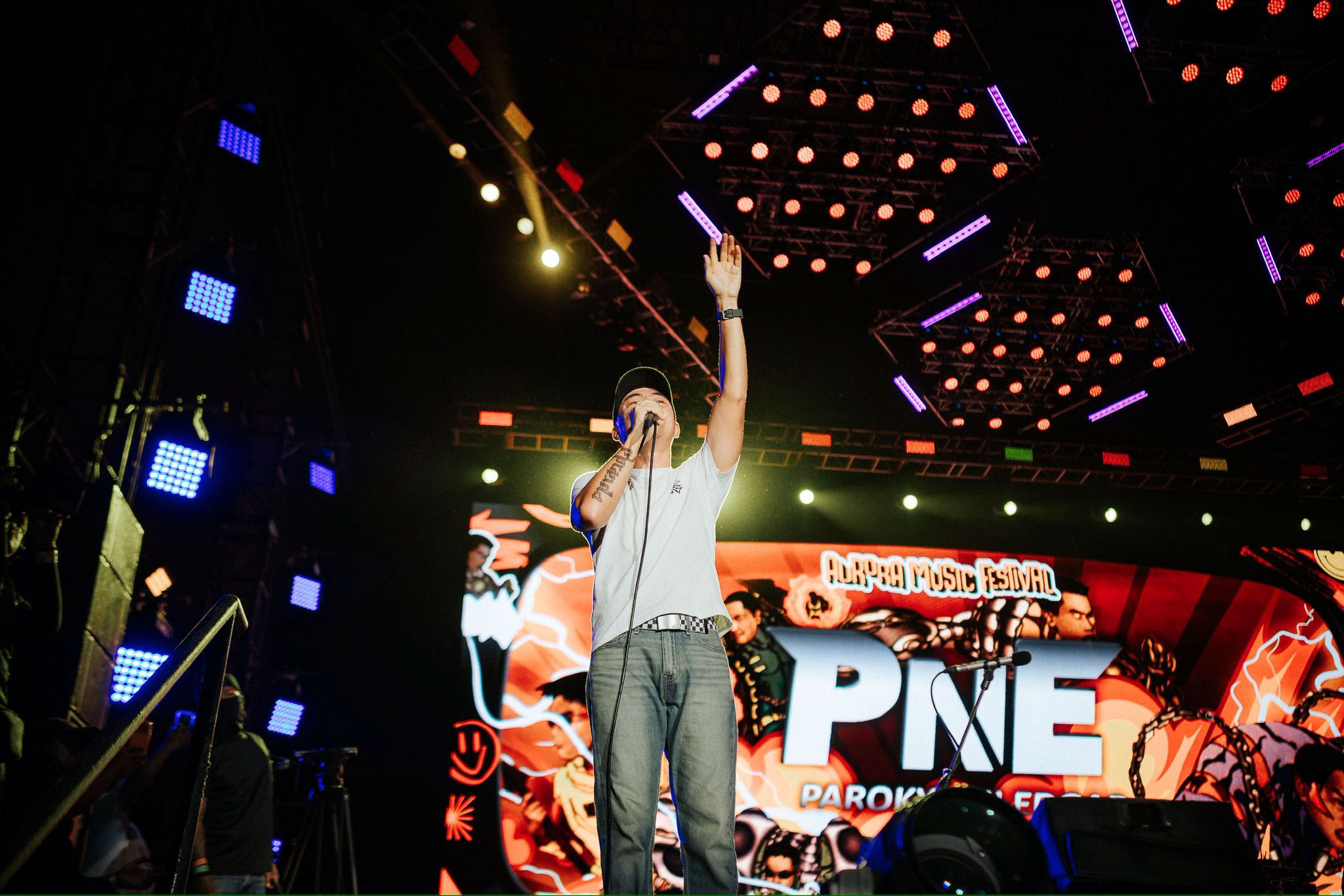Male singer on stage, raising his hand while performing at a colorful outdoor music festival, with bright red and purple lights overhead.