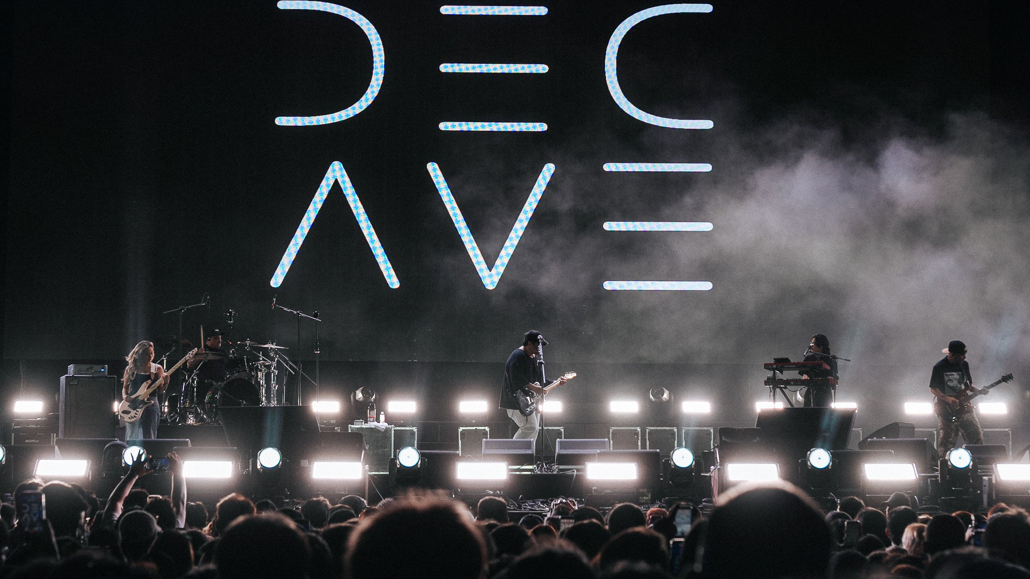 Live concert scene: a band performs on stage with guitars and keyboards, large illuminated backdrop, and a cheering crowd in the foreground.