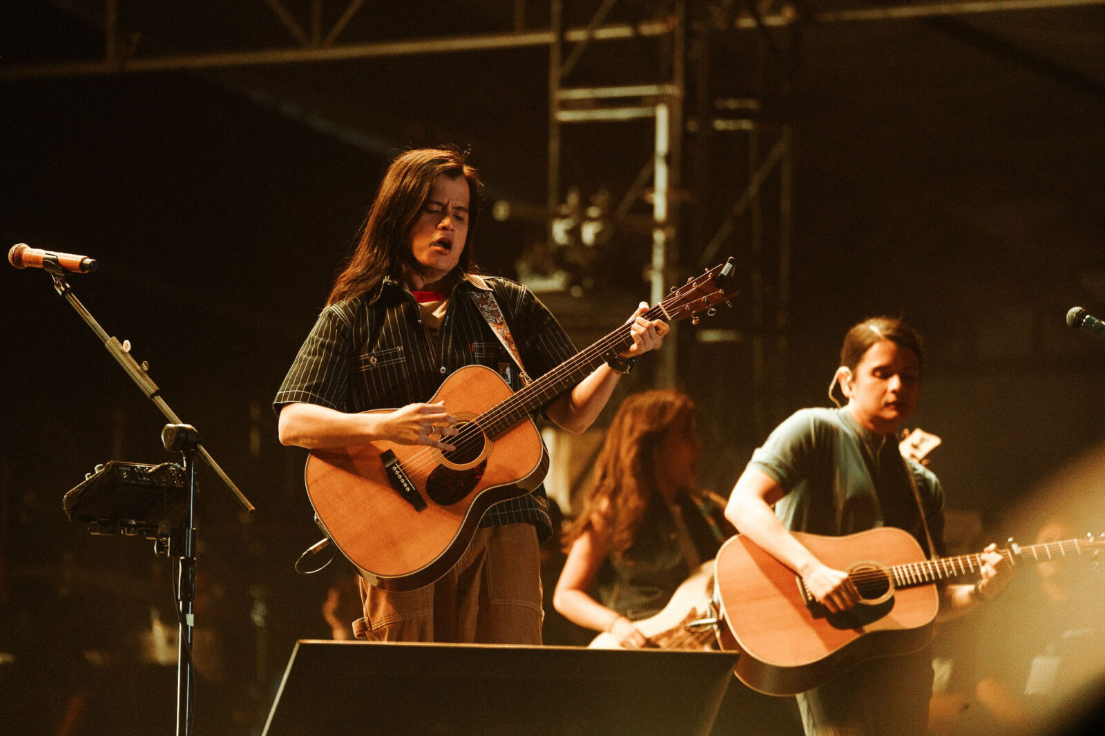 Female musician center stage singing and playing an acoustic guitar during a live concert, with bandmates in the background on stage.