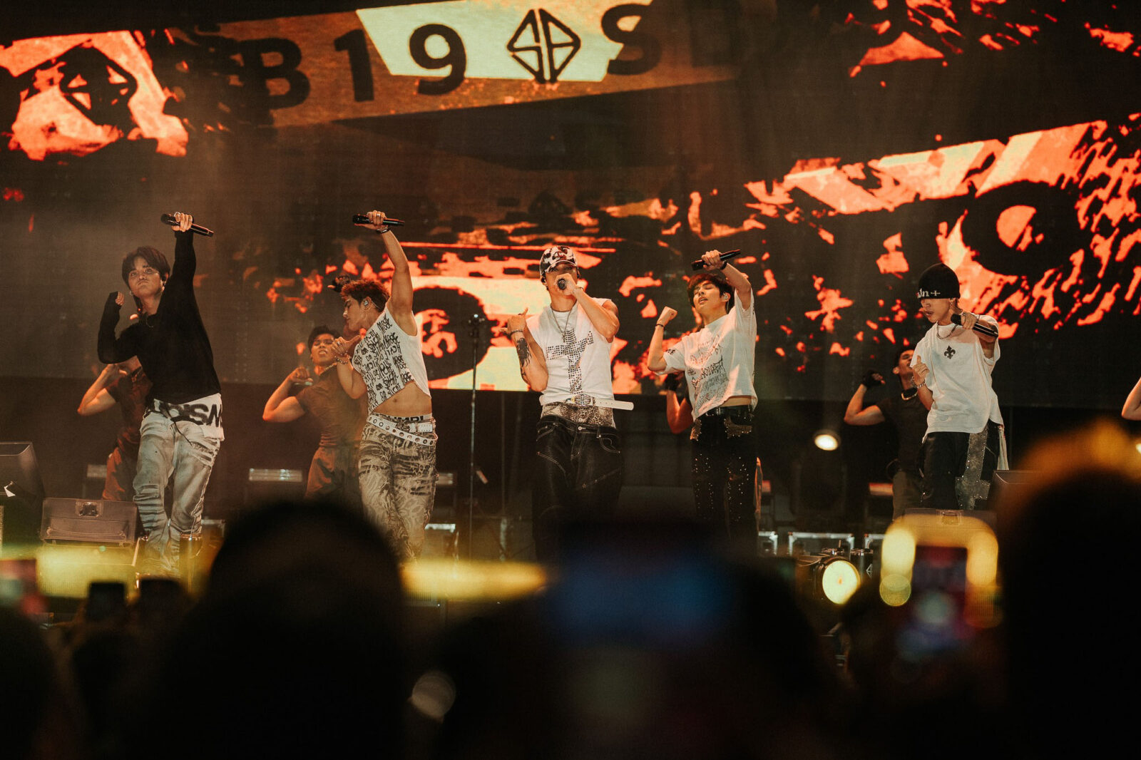 Group of male performers on stage singing into microphones during a concert, with a fiery red backdrop and cheering crowd in the foreground.