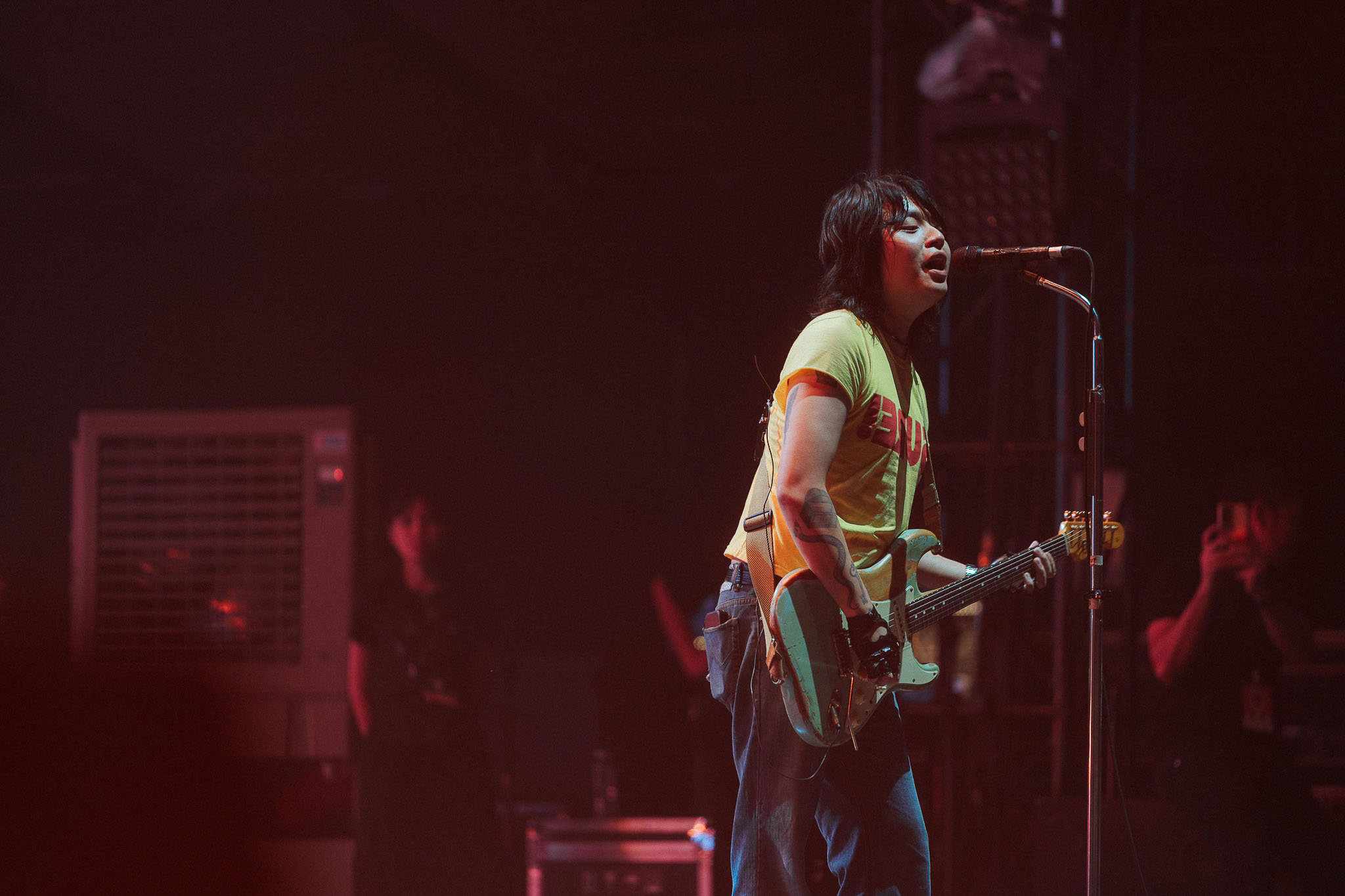 Female guitarist singing into a microphone on stage with a turquoise electric guitar under red stage lighting.