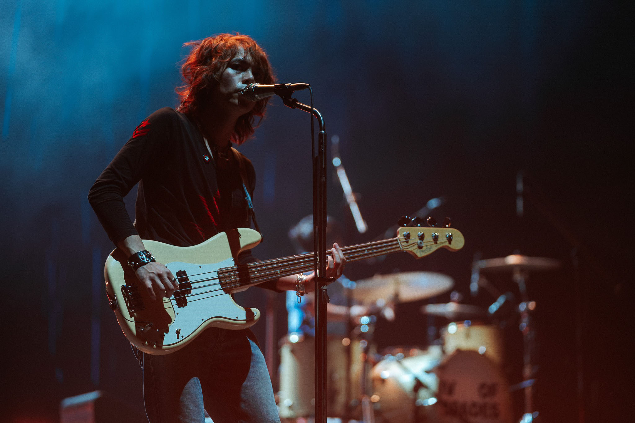 Bass guitarist sings into a microphone while playing a cream-colored bass on a dimly lit concert stage with blue lighting in the background.