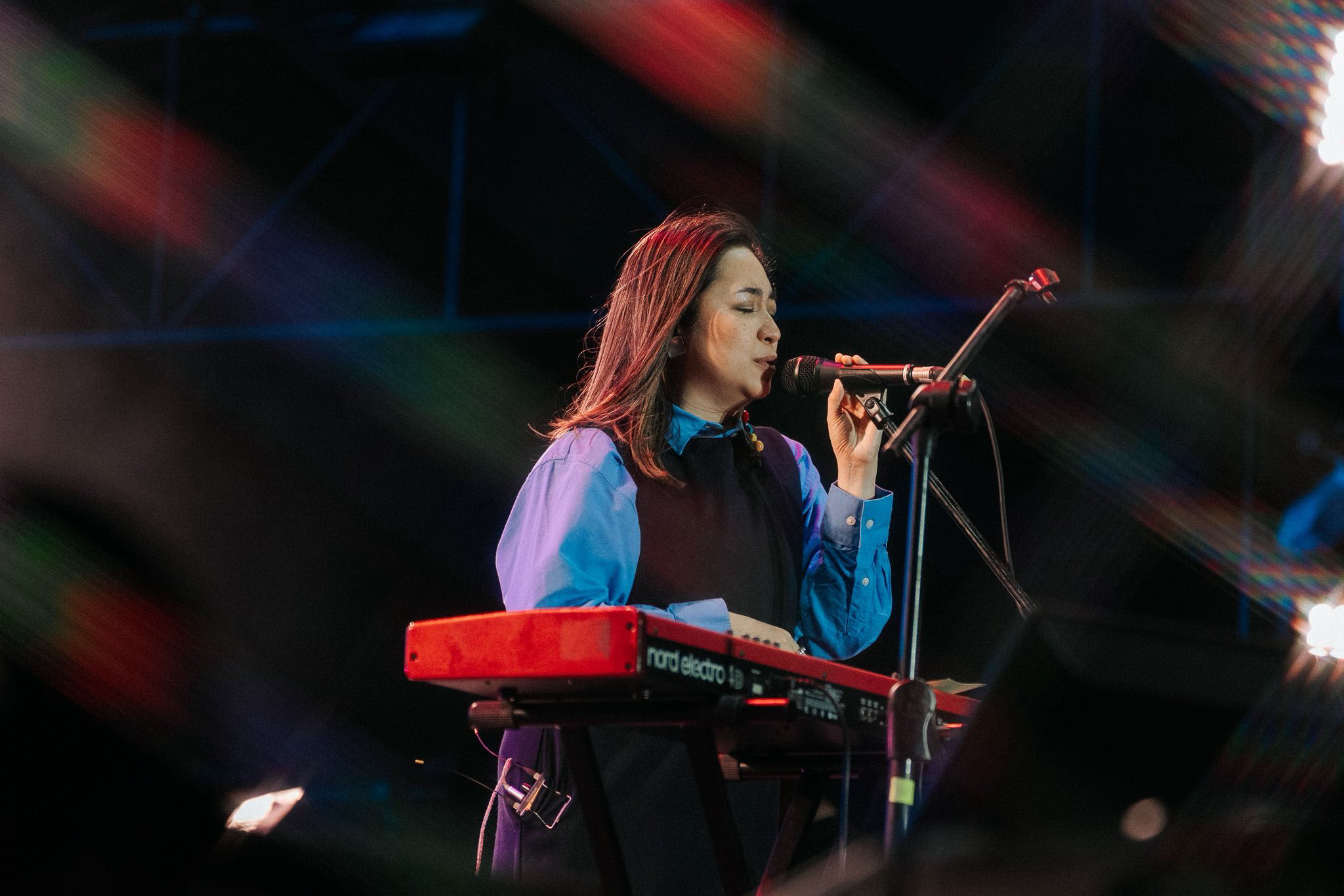 Female singer at a keyboard on stage, singing into a microphone with colorful stage lights behind her.