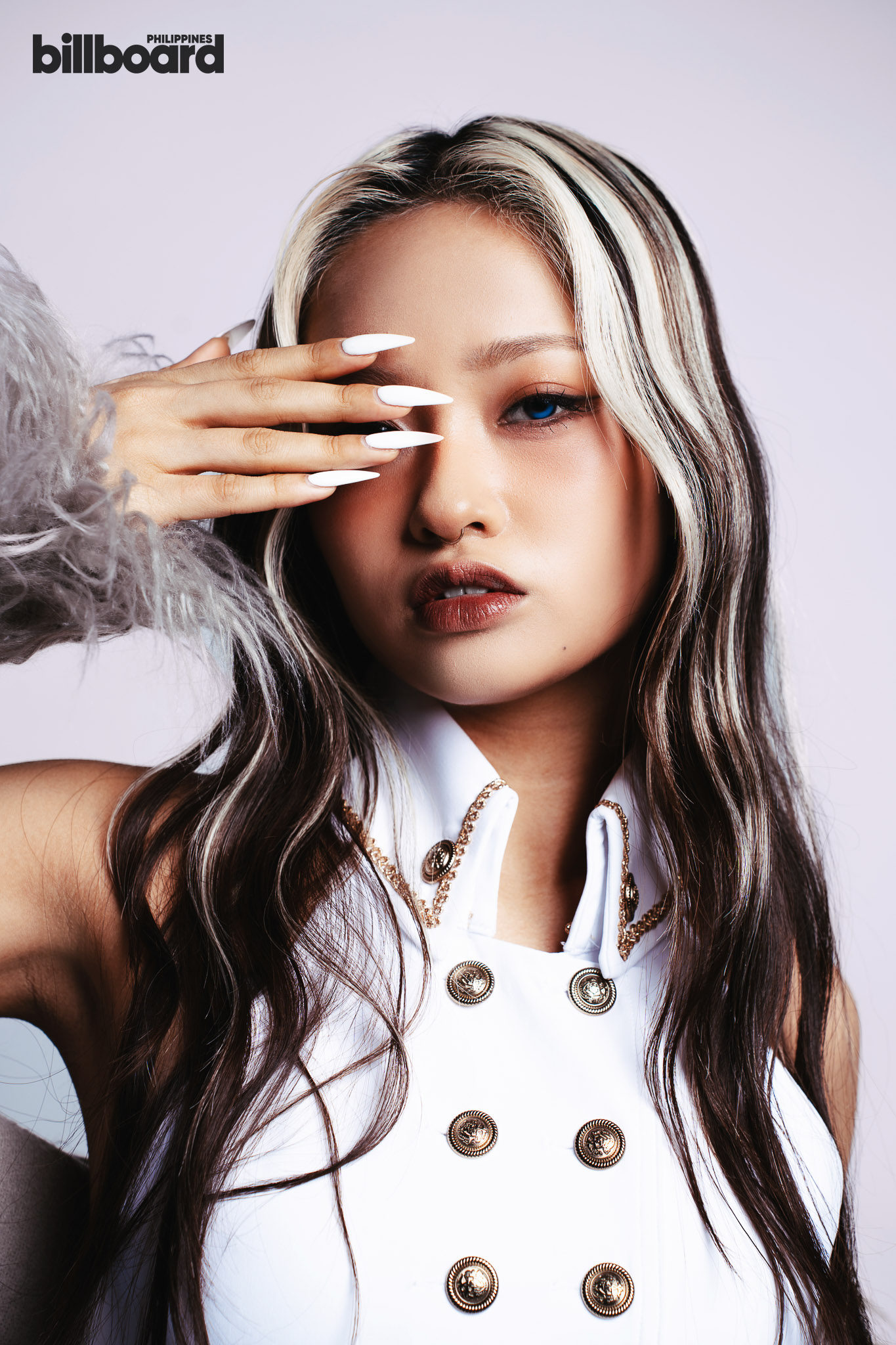 Portrait of a woman with highlighted hair covering one eye with long white nails, wearing a white sleeveless top with gold buttons; billboard philippines logo in top-left.