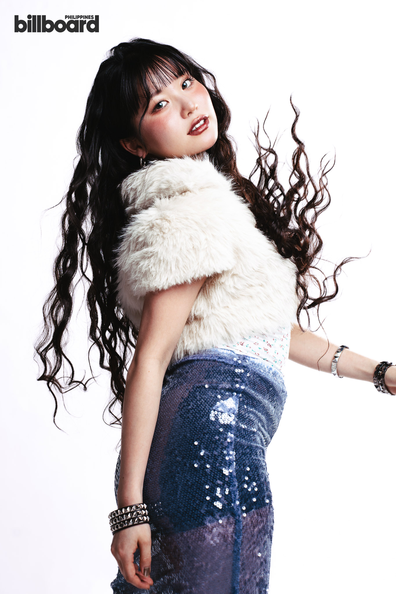 Portrait of a young woman with long dark curls wearing a white faux-fur vest and a blue sequined skirt, looking over her shoulder.