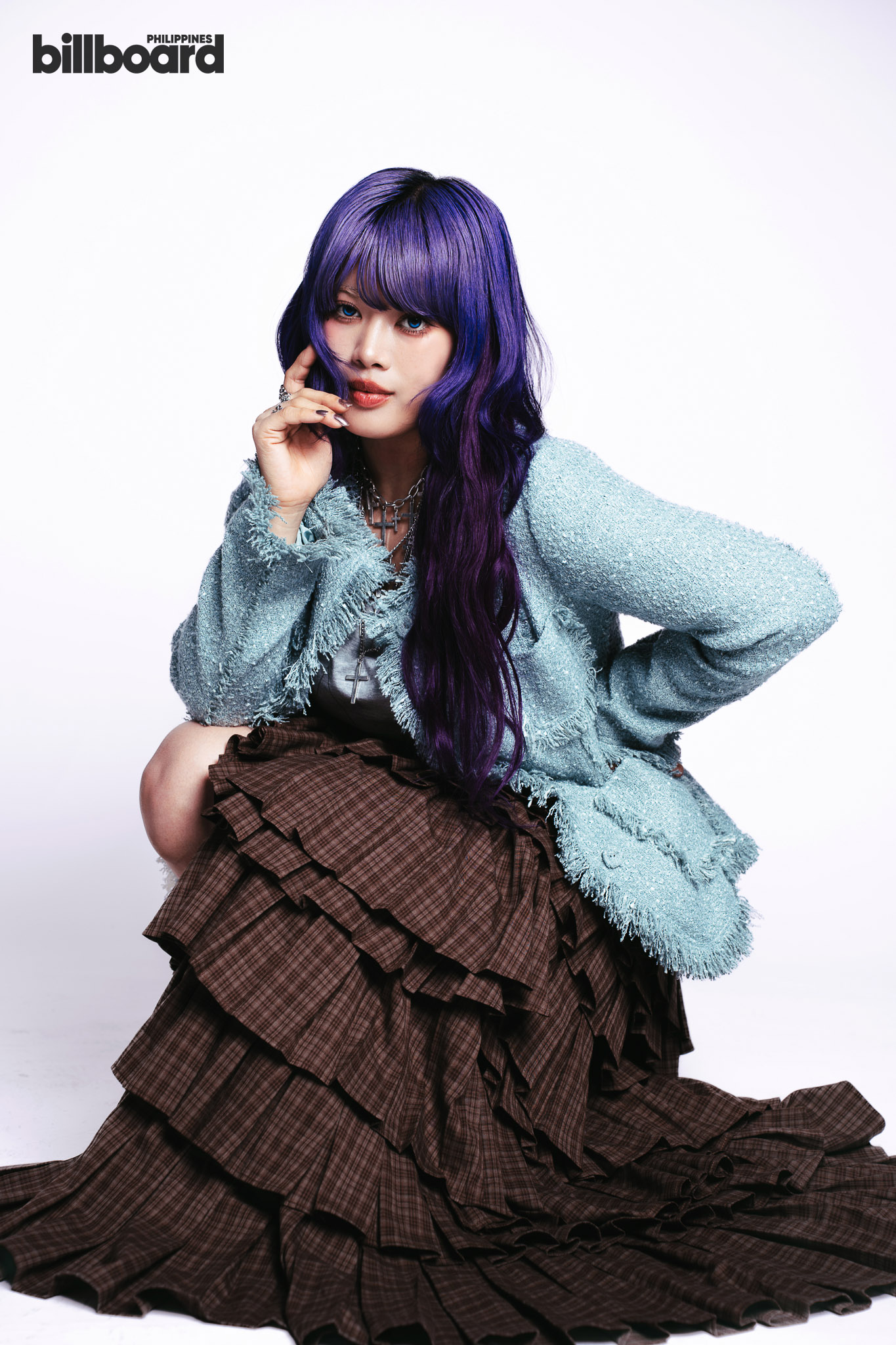 Portrait of a woman with purple hair wearing a light blue textured jacket and a layered brown skirt, posing with her hand near her face; billboard logo visible in the top-left corner.