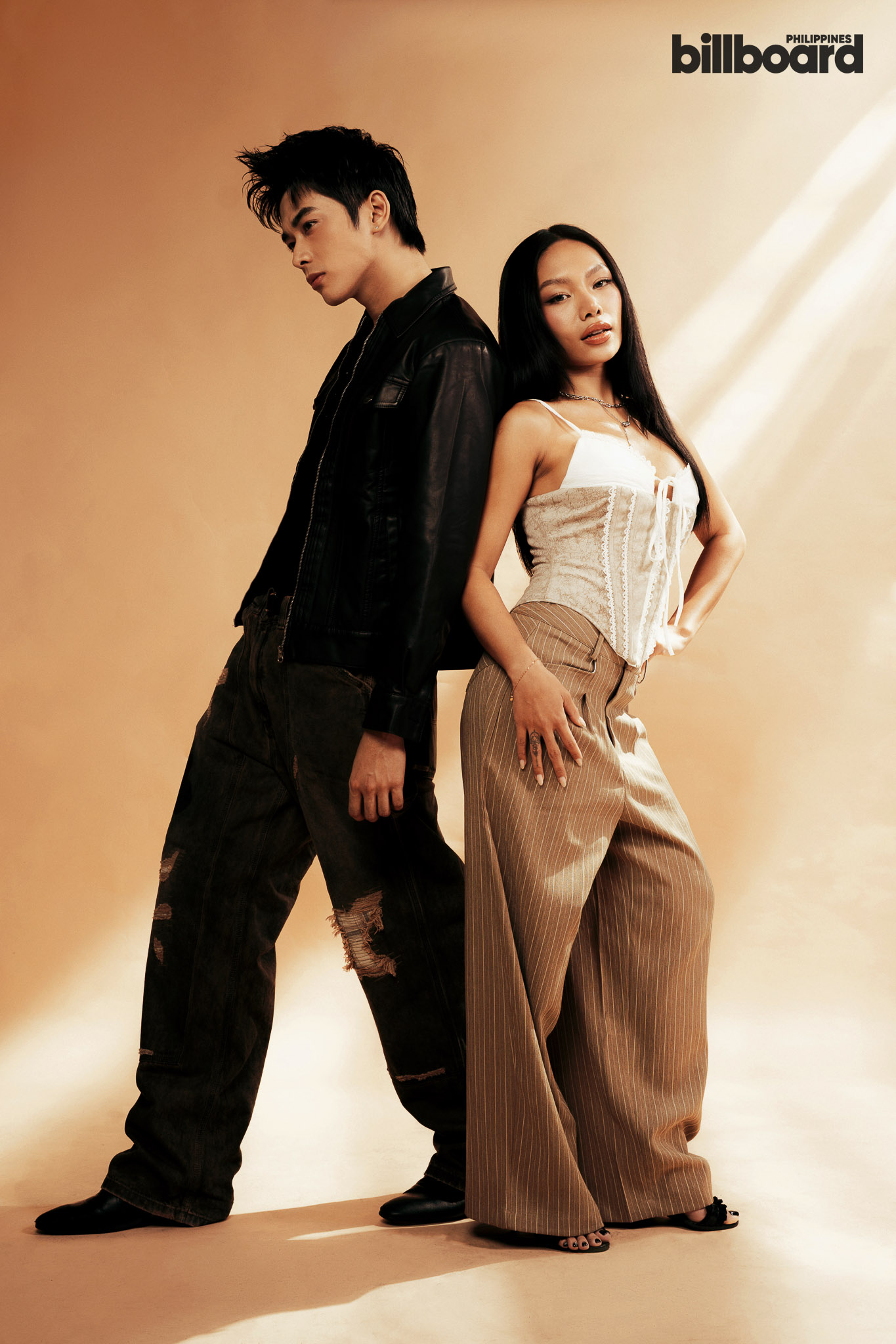 Two models back-to-back in a warm studio backdrop; man in black leather jacket and ripped jeans, woman in white corset and striped pants.