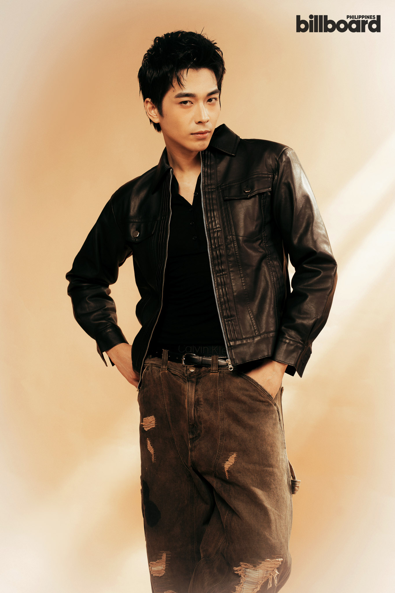 Male model wearing a black leather jacket and dark shirt, posing with hands in pockets against a warm beige gradient background.
