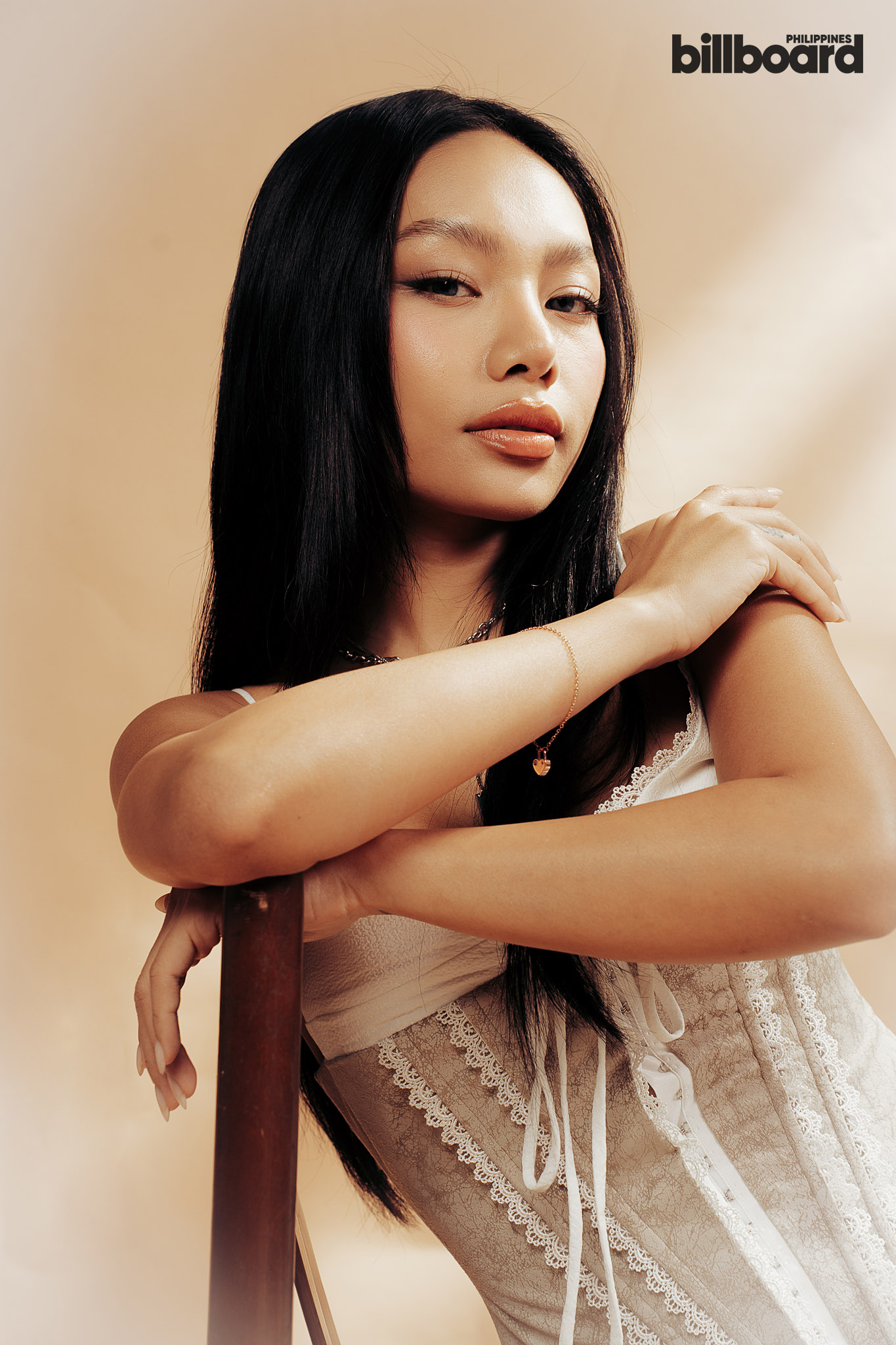 Portrait of a woman with long black hair wearing a beige lace dress, resting her arms on a chair, soft beige backdrop, Billboard Philippines logo in the corner.