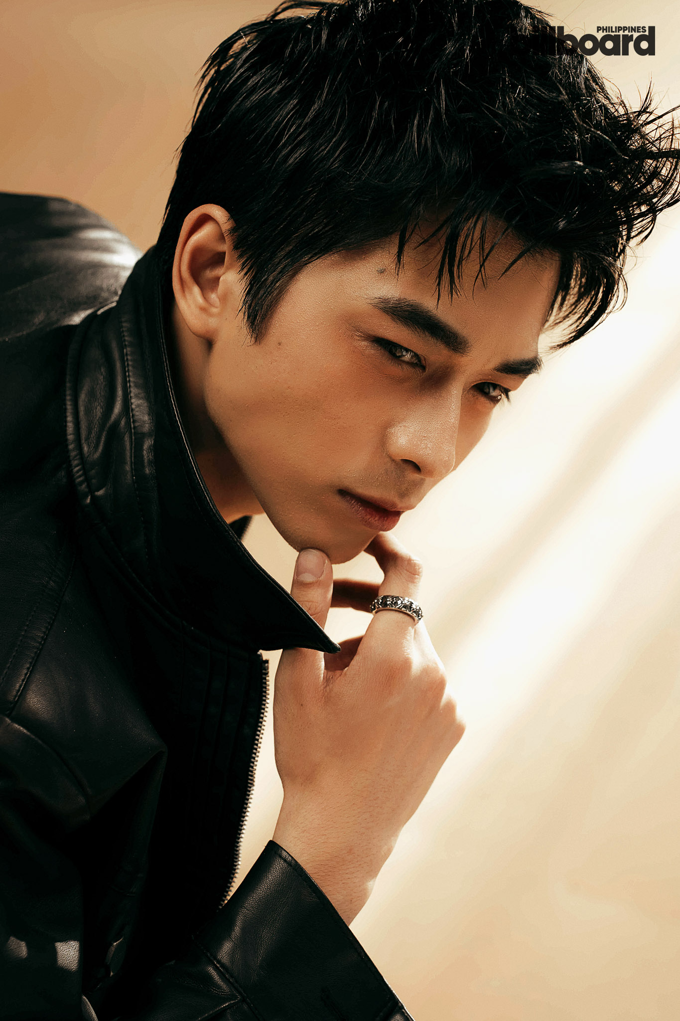 Close-up of a man in a black leather jacket looking downward, hand near his chin with rings, in a studio setting.