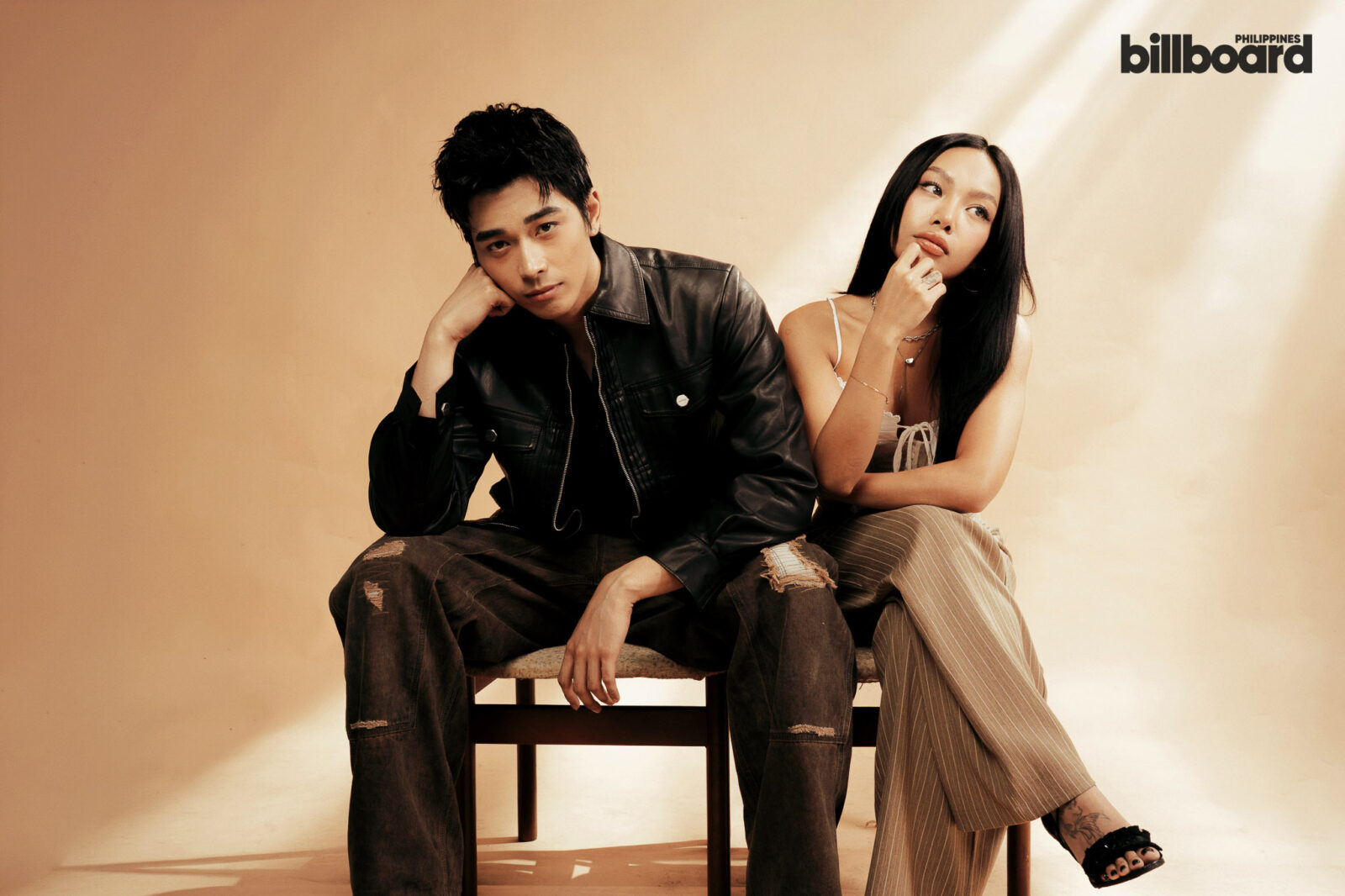Two young adults sit closely on a bench against a warm beige studio backdrop; man wears a black leather jacket, ripped jeans, woman wears striped pants and a white top, Billboard Philippines logo in the top-right corner.