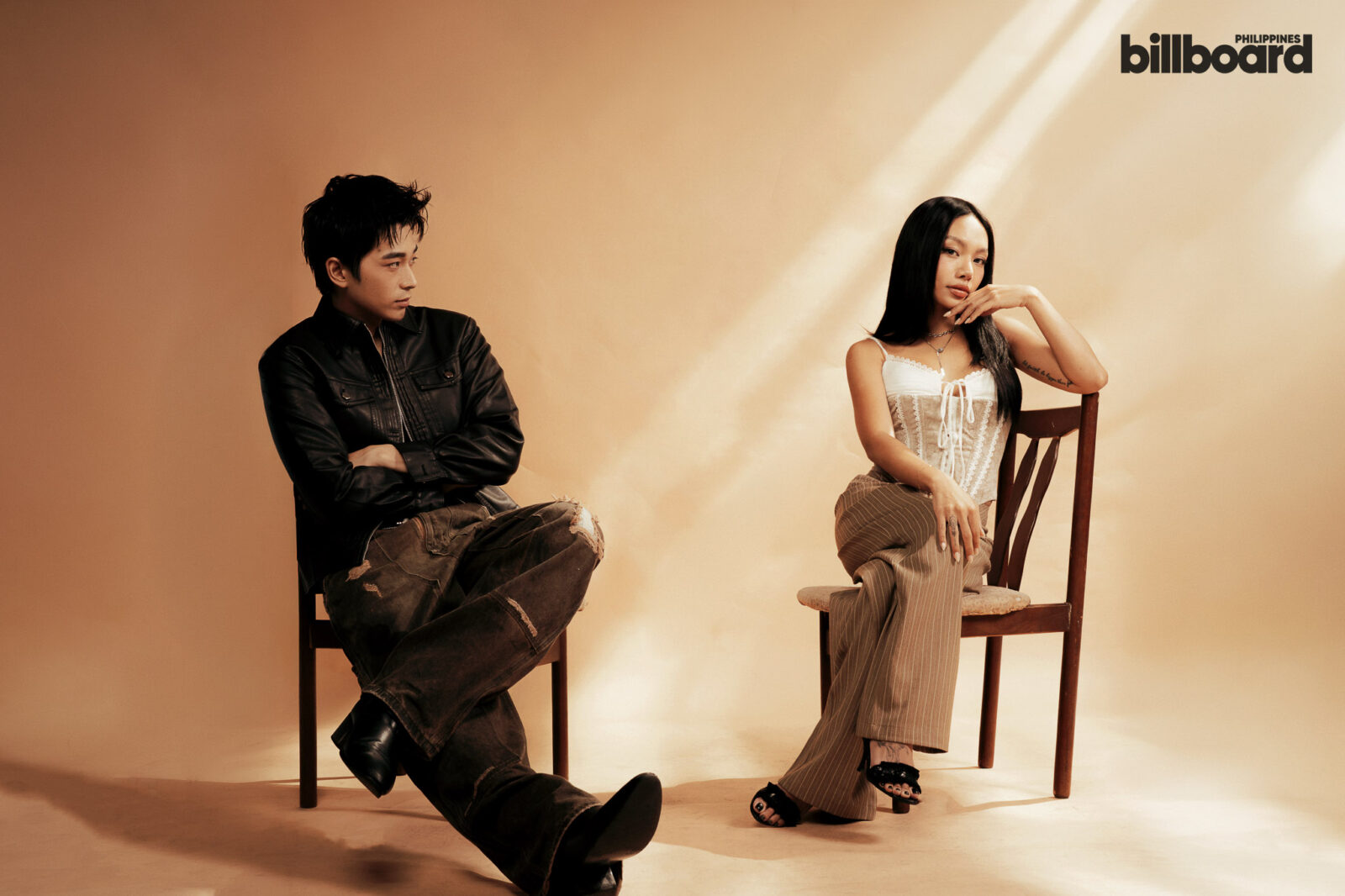 Two people sit on chairs against a warm beige studio backdrop; a man in a black jacket looks toward a woman in a white corset top on the right.