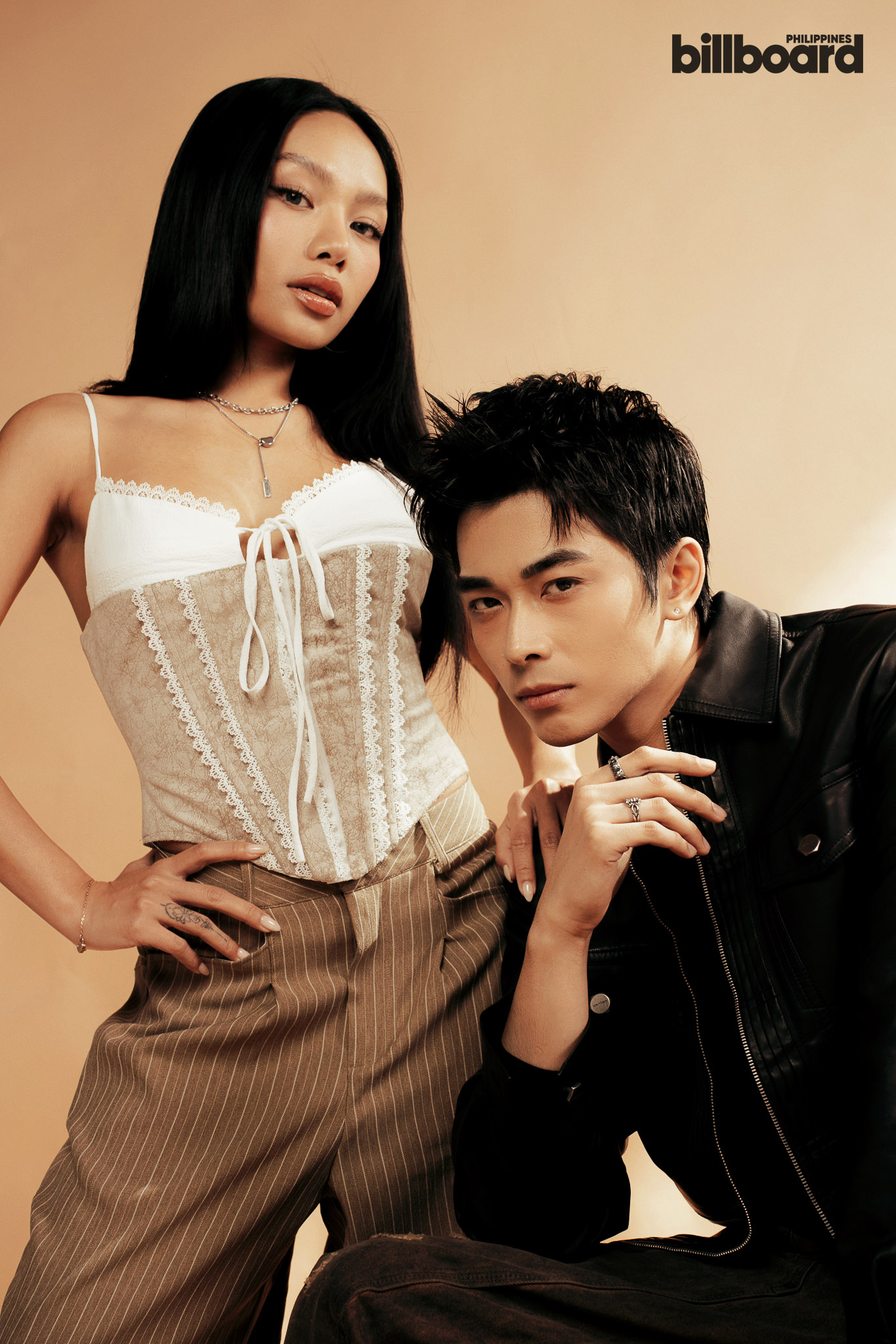 Two models pose stylishly against a warm tan backdrop, woman in a white lace corset and striped pants, man in a black leather jacket beside her. Billboard Philippines logo in the corner.