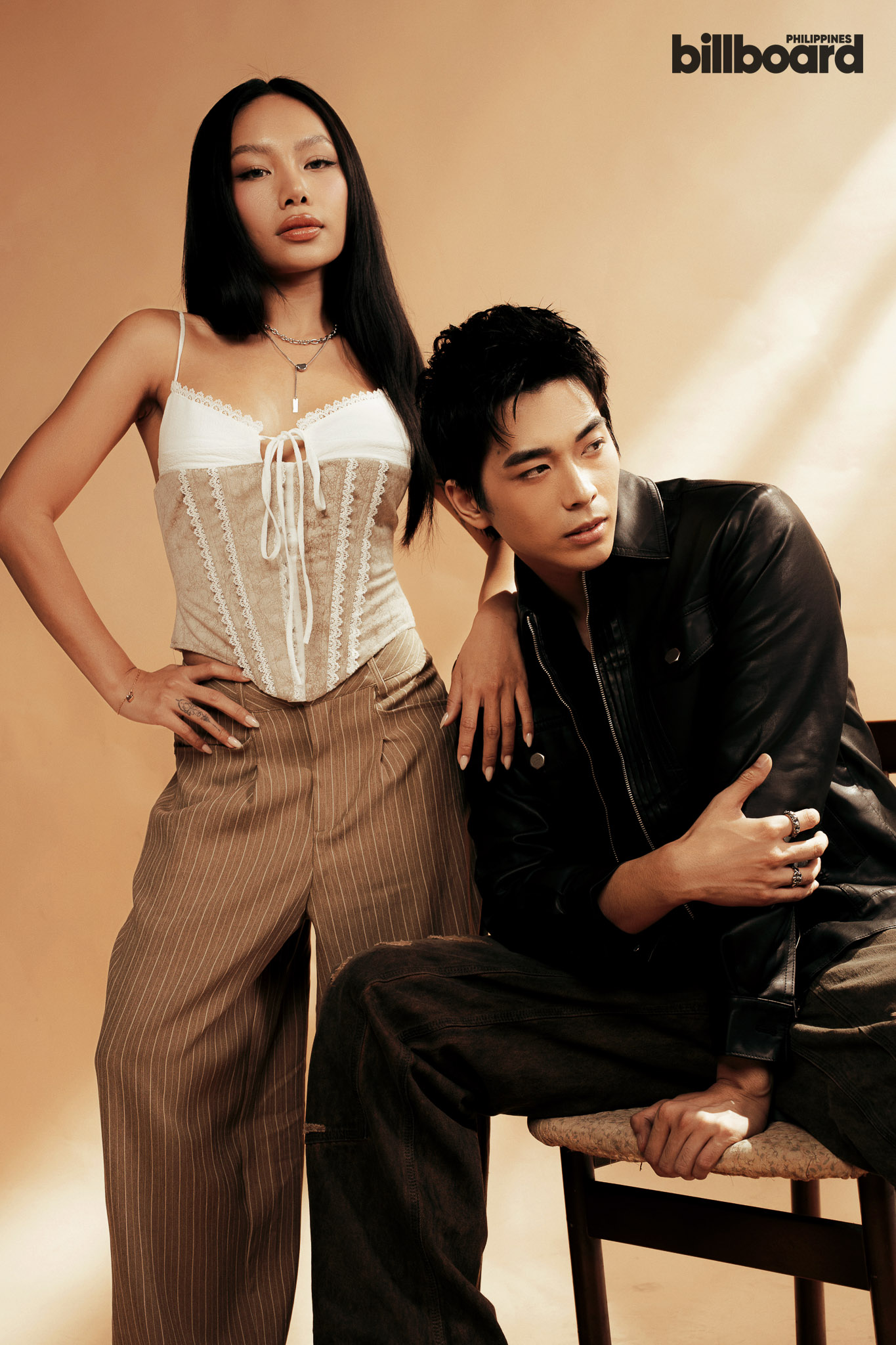 Studio fashion shot: woman in a white lace corset and pinstripe pants stands with hand on hip beside a man in a black leather jacket; Billboard logo top right.