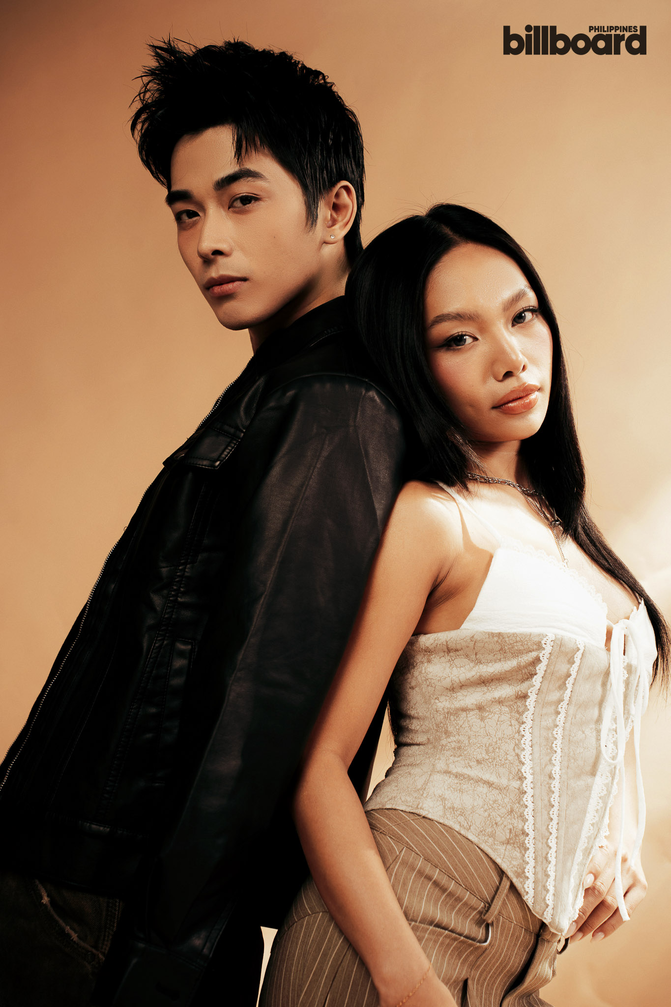 Two models lean back-to-back against a warm beige backdrop; man in a black leather jacket and woman in a lace corset top, Billboard Philippines logo in the corner.