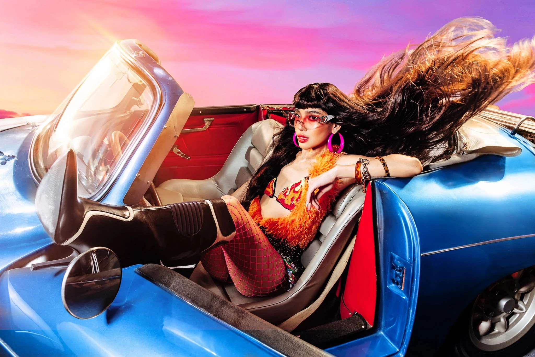 Woman in an orange fur outfit and pink accessories sits in a blue convertible as hair whips in the wind against a colorful sunset sky.