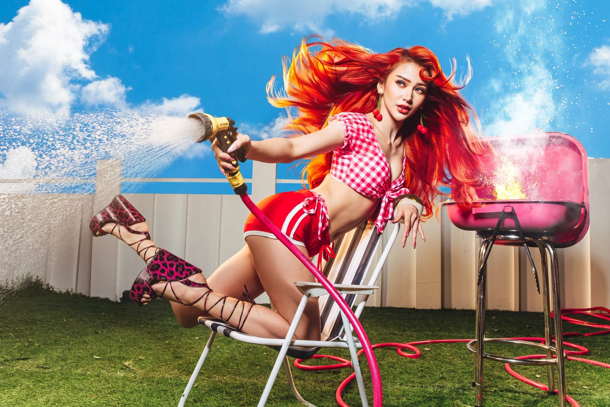 Woman with long red hair in a pink checkered bikini sprays water from a hose toward a pink barbecue grill in a sunny backyard.