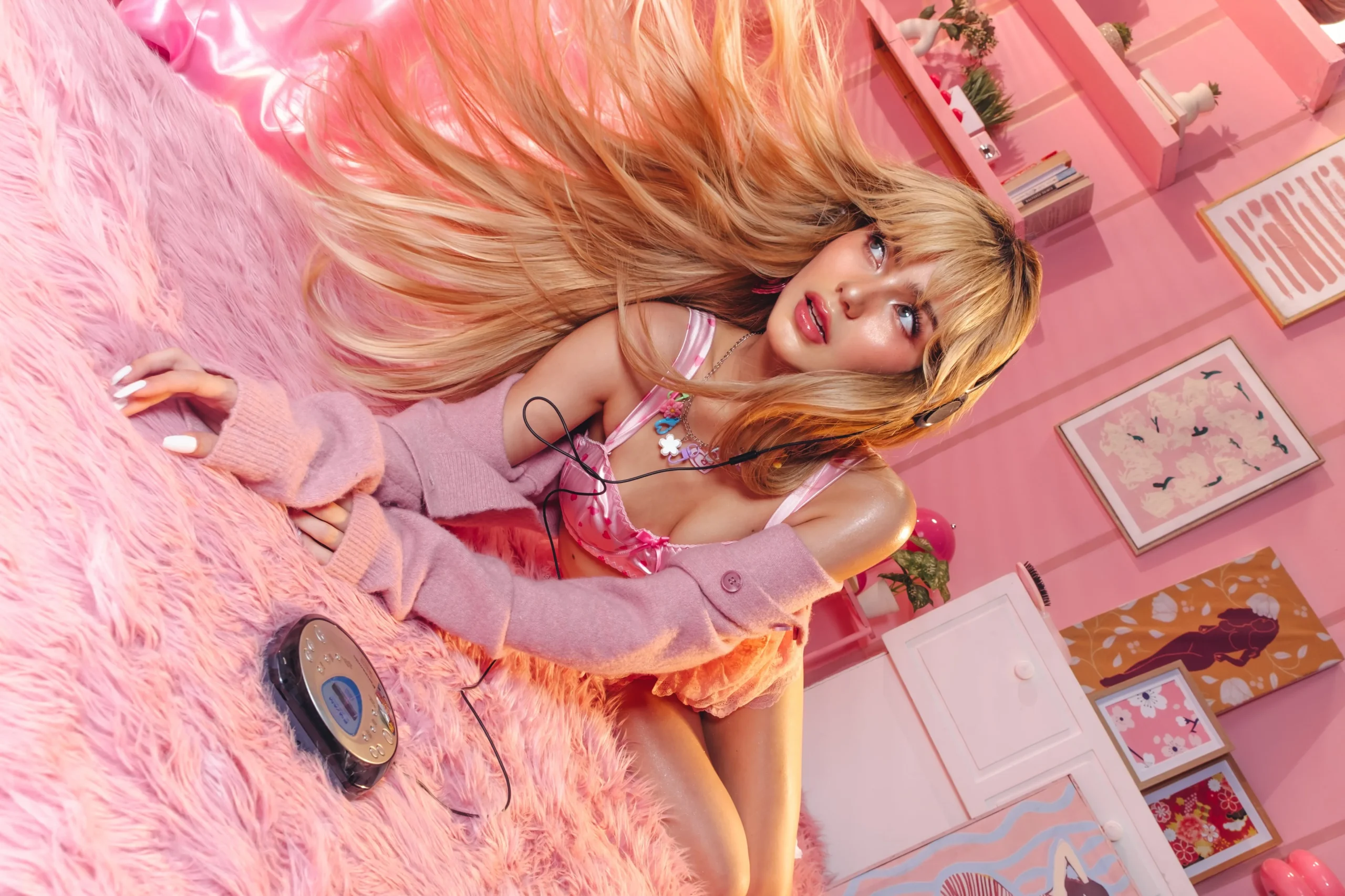 Woman with long blonde hair lounging on a pink fluffy rug in a pink-themed room, wearing a pink top and headphones.